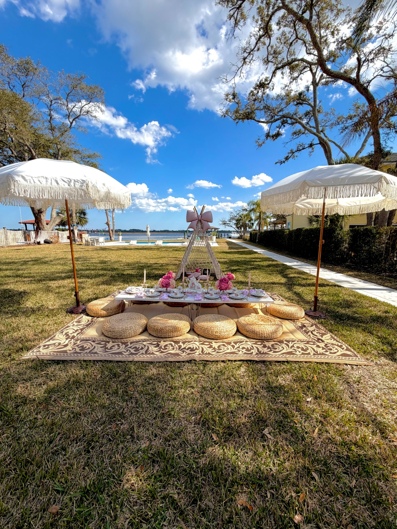 Outdoor setup with cushions, umbrellas, and a table with decorations near water, under a blue sky with clouds.