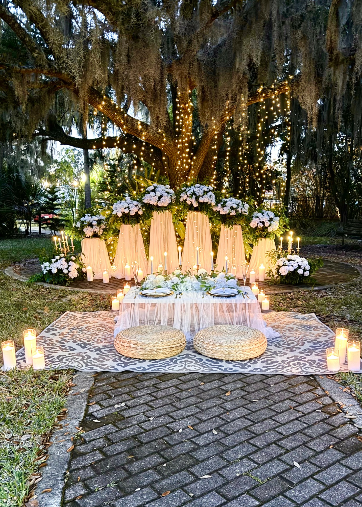 Outdoor setup with illuminated tree, floral arrangements, candles, and cushions on a patterned rug on cobblestone ground.