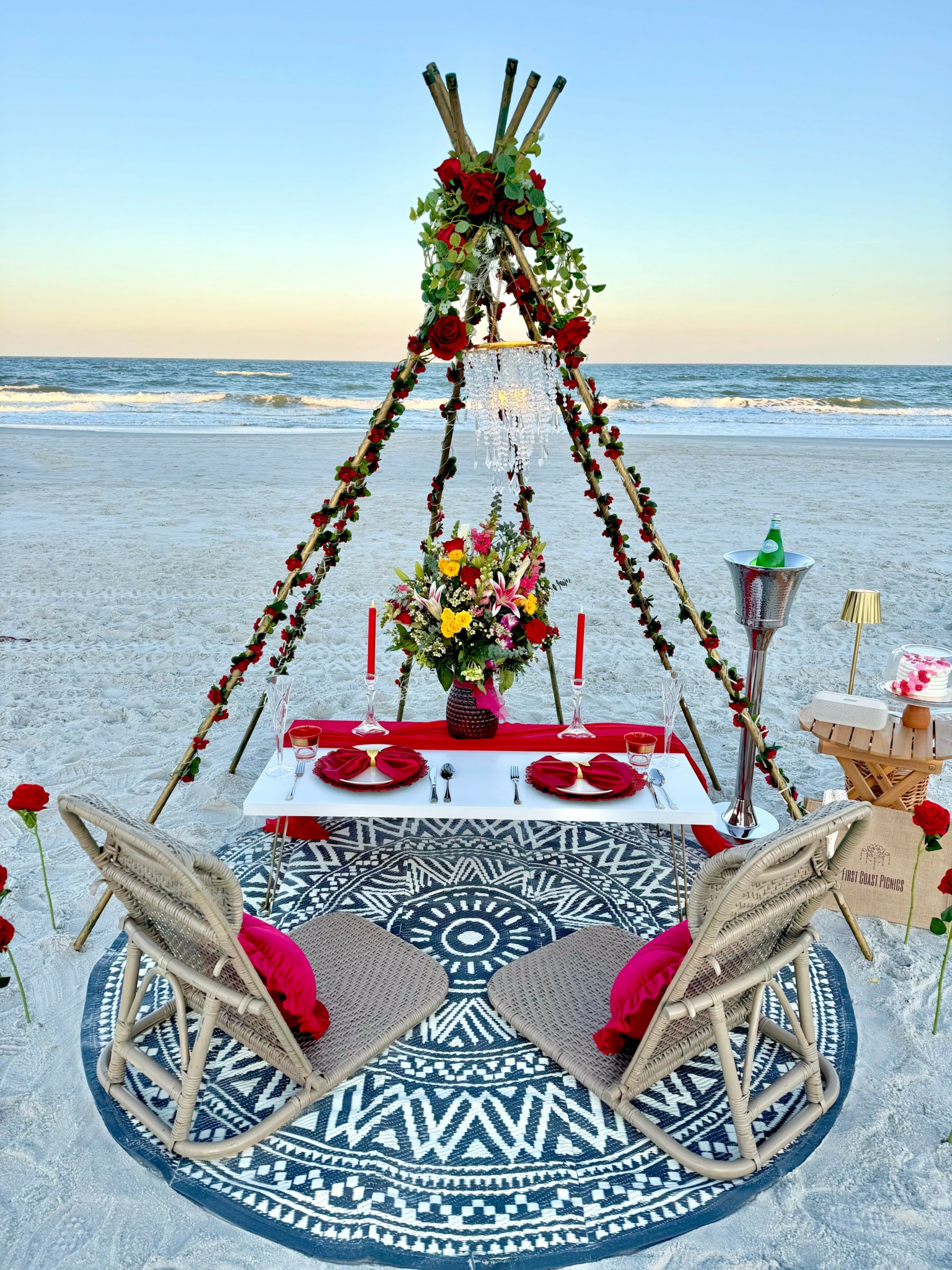 Beach setup with a floral teepee, table, and chairs for dining, overlooking the ocean at sunset.