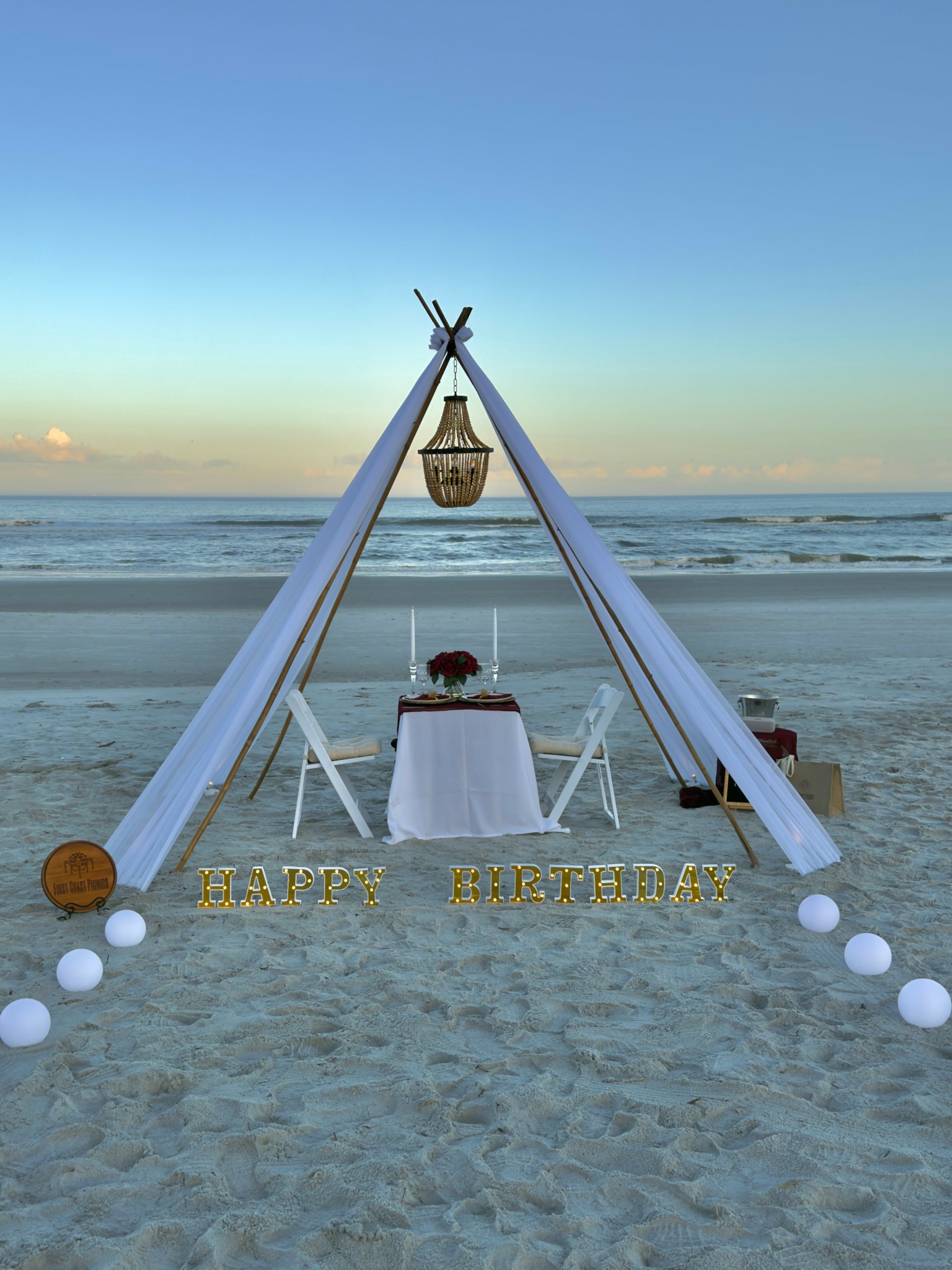 Beach setup with a teepee structure, table with flowers, and decorative signs reading 