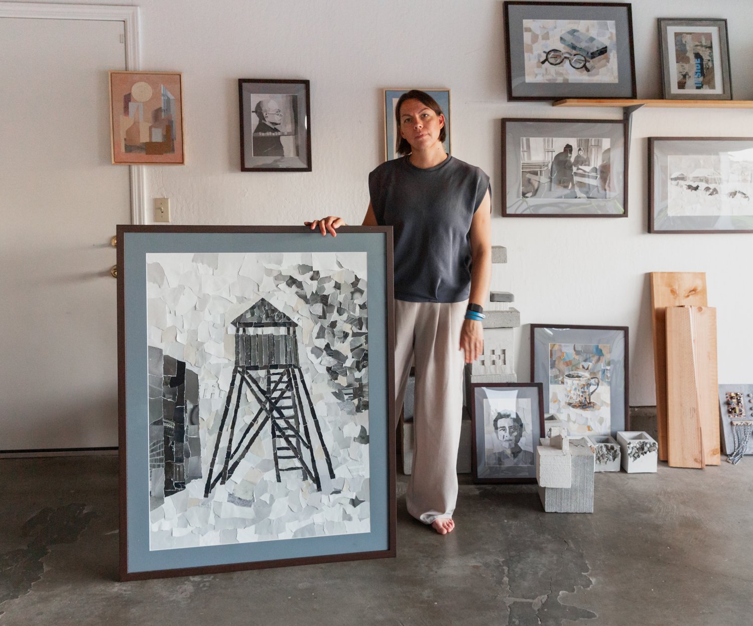 Woman standing next to a large framed artwork of a watchtower, surrounded by other framed artworks on the wall.