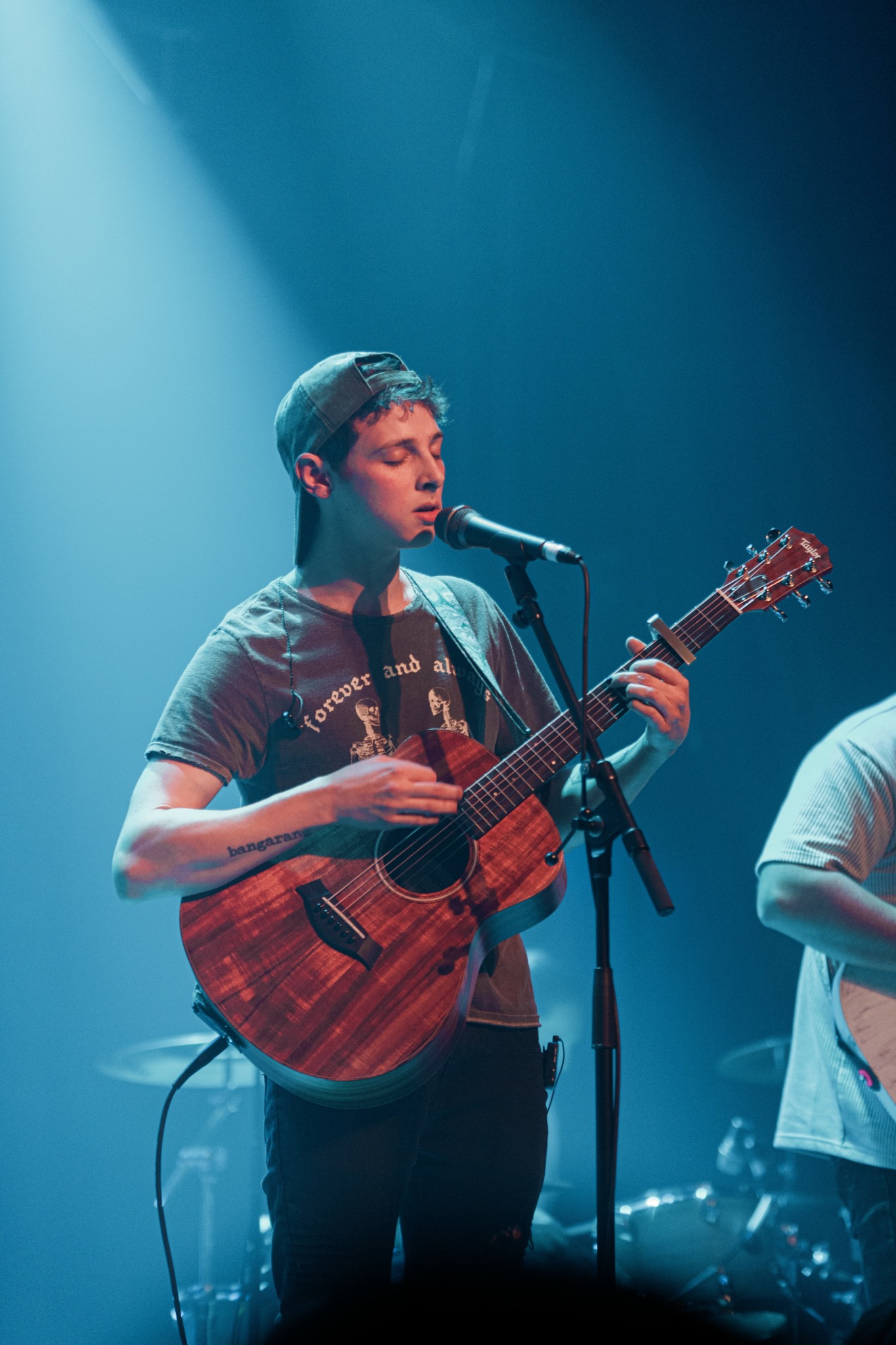 Young person with short hair singing into a microphone while playing an acoustic guitar on stage.