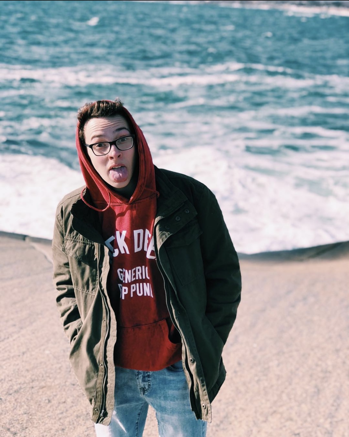 Person with glasses sticking out tongue, wearing a red hoodie and green jacket, standing on beach near water.