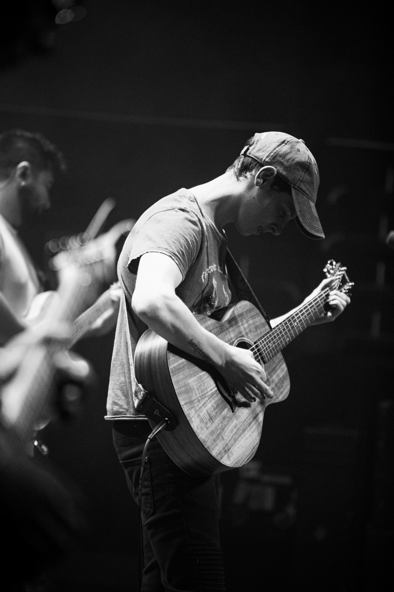 Person playing an acoustic guitar, wearing a cap, in a dark setting with blurred background.