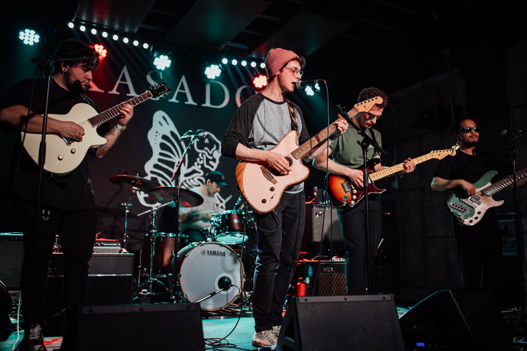 Five musicians perform on stage with guitars and drums, with a butterfly and text in the background.