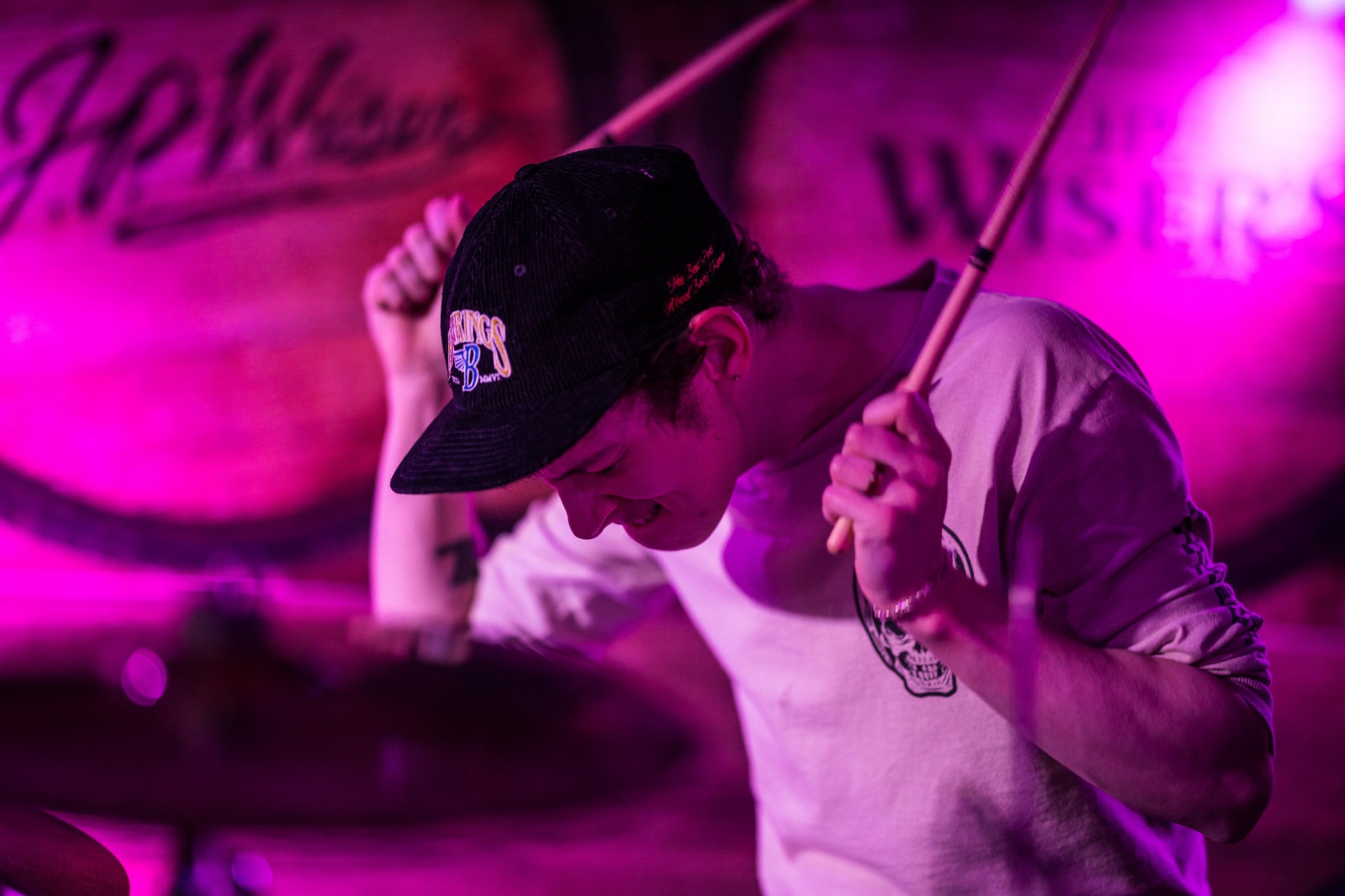 Person wearing a black cap playing drums in a pink-lit setting.