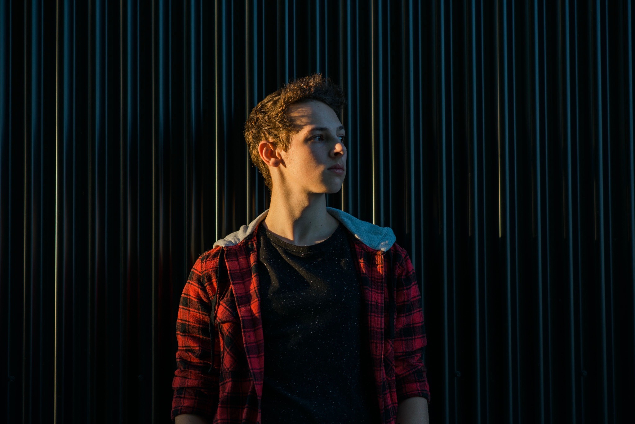 Young person with short hair wearing a red and black checkered shirt standing against a dark, vertically striped background.