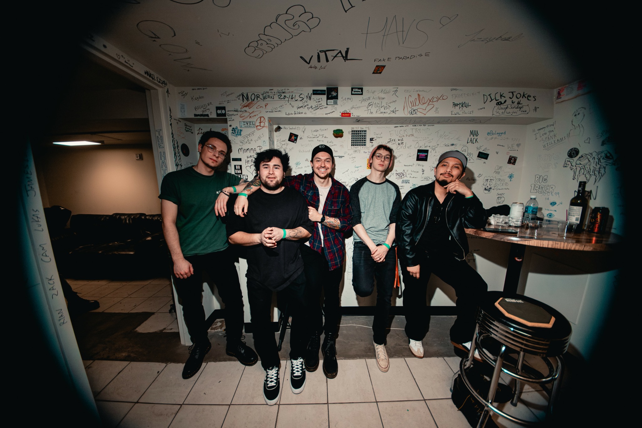 Five men standing together in a room with a wall covered in drawings and notes, some holding drinks.