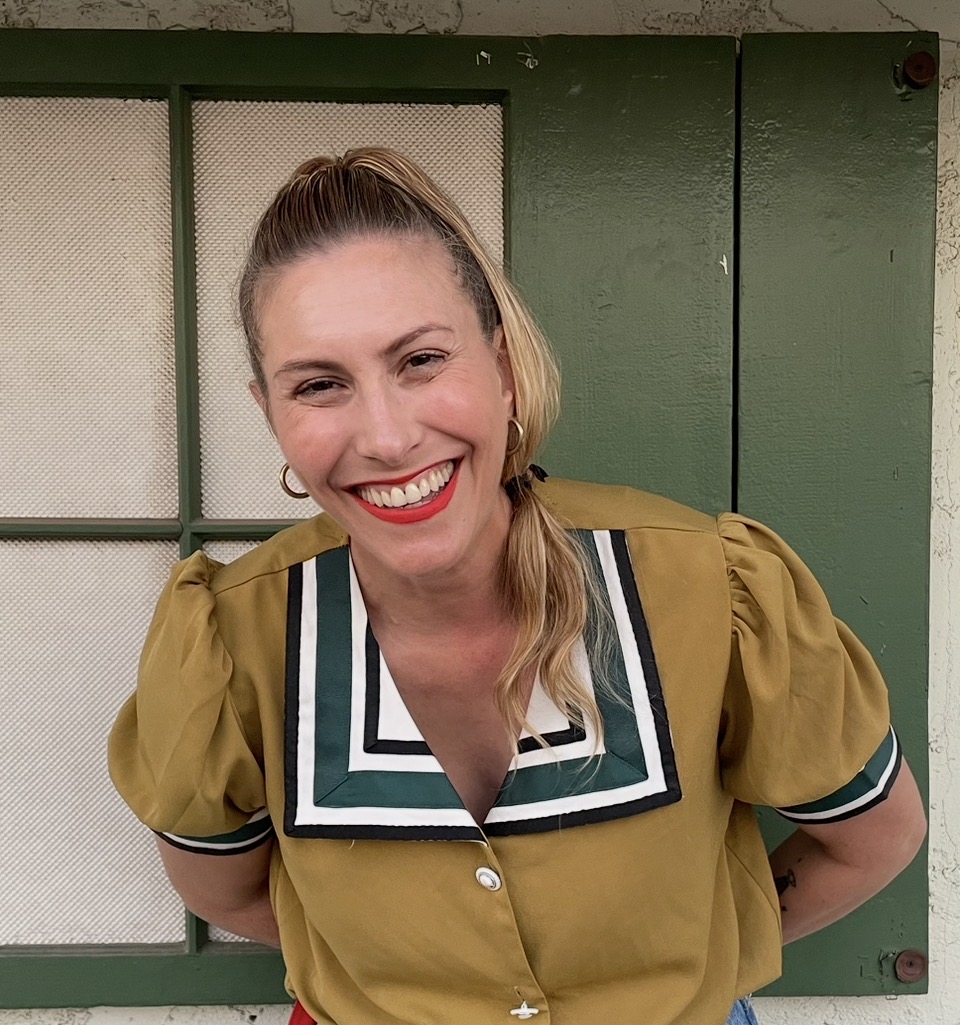 Woman with blonde hair in a ponytail, smiling, wearing a mustard yellow top with a large collar, standing in front of a green door.