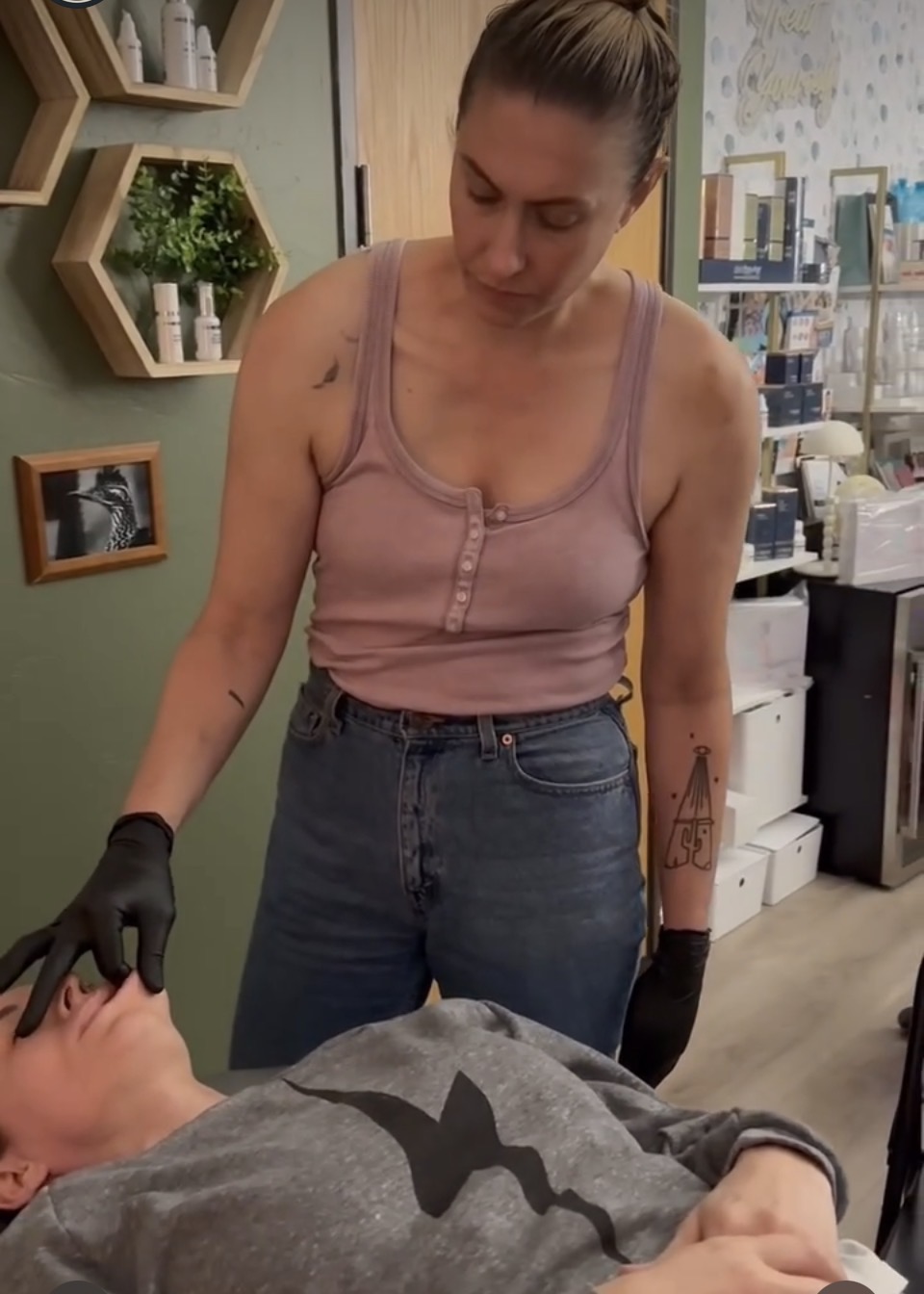 Woman standing over a person lying down, wearing a pink tank top and jeans, in a room with shelves and equipment.