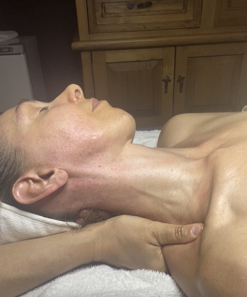 Person lying down with head tilted back, receiving a massage on neck and shoulder area, in a room with wooden furniture.