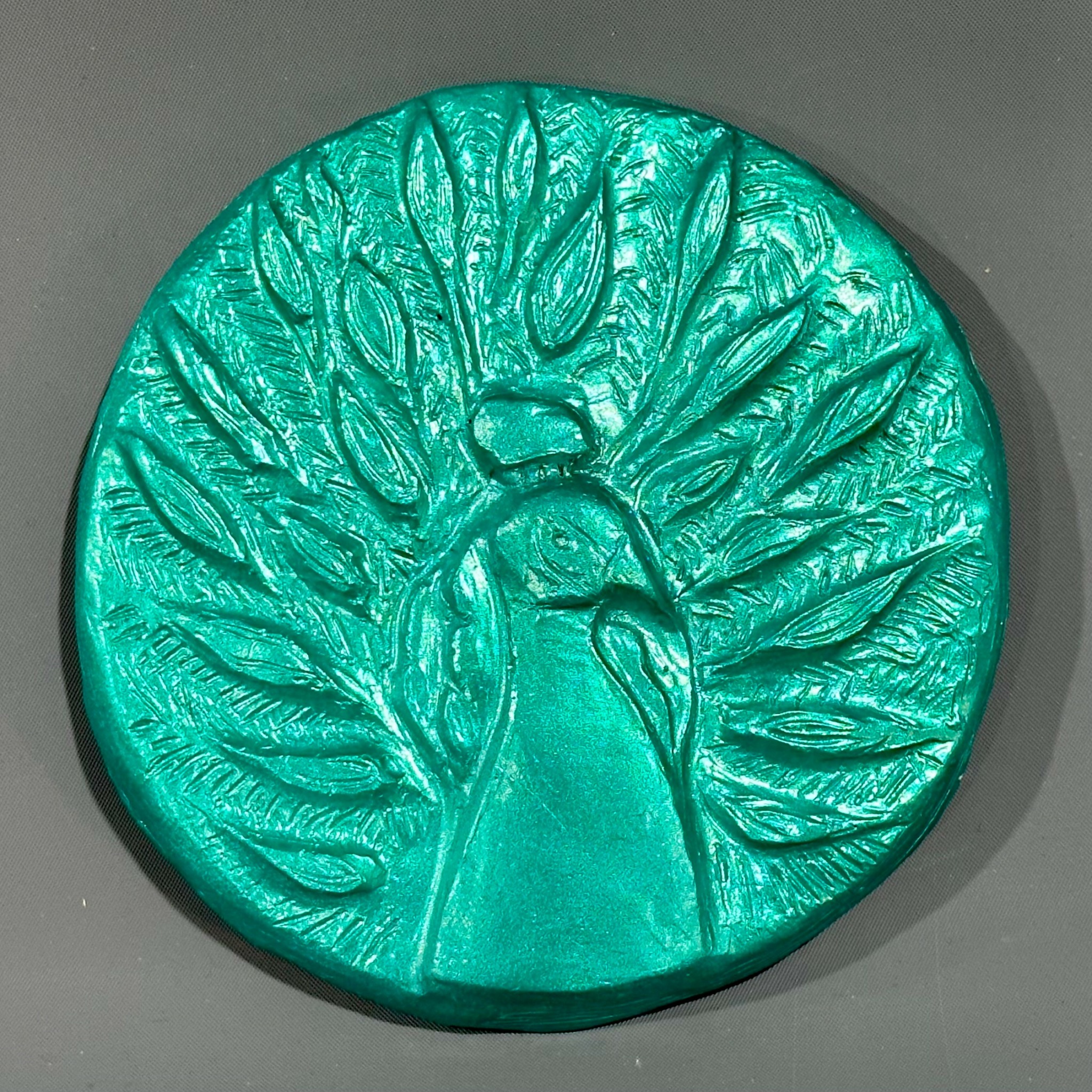 Round teal-colored relief sculpture of a peacock with spread tail feathers.