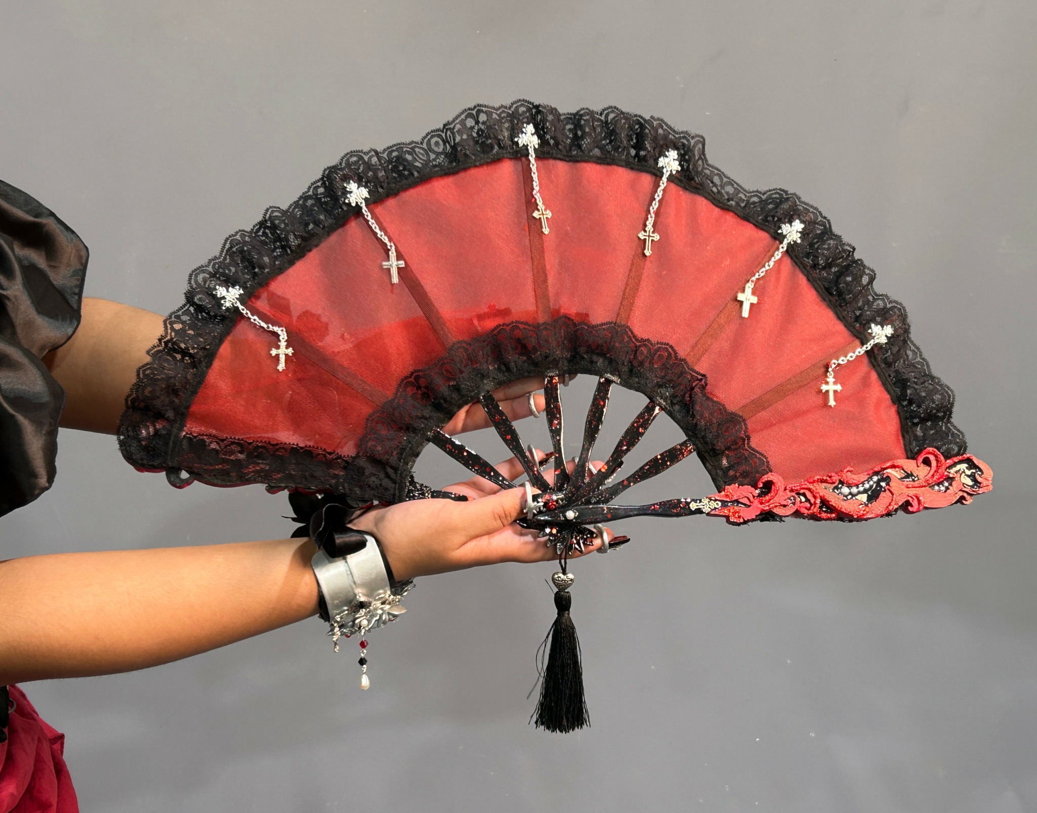 Person holding a decorative fan with black lace, red fabric, and small cross charms, against a gray background.