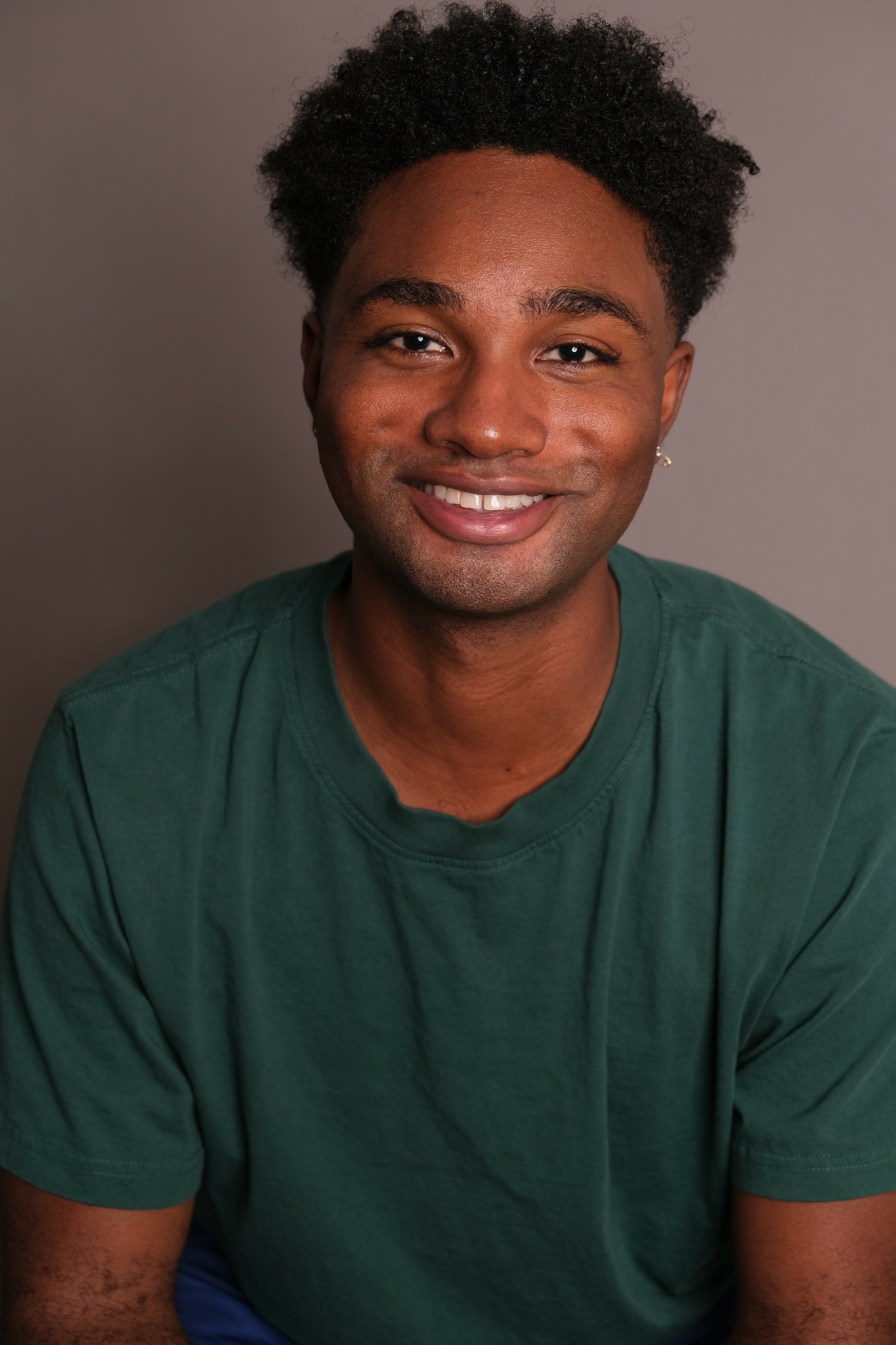 Young man with curly hair smiling, wearing a green T-shirt, against a plain background.