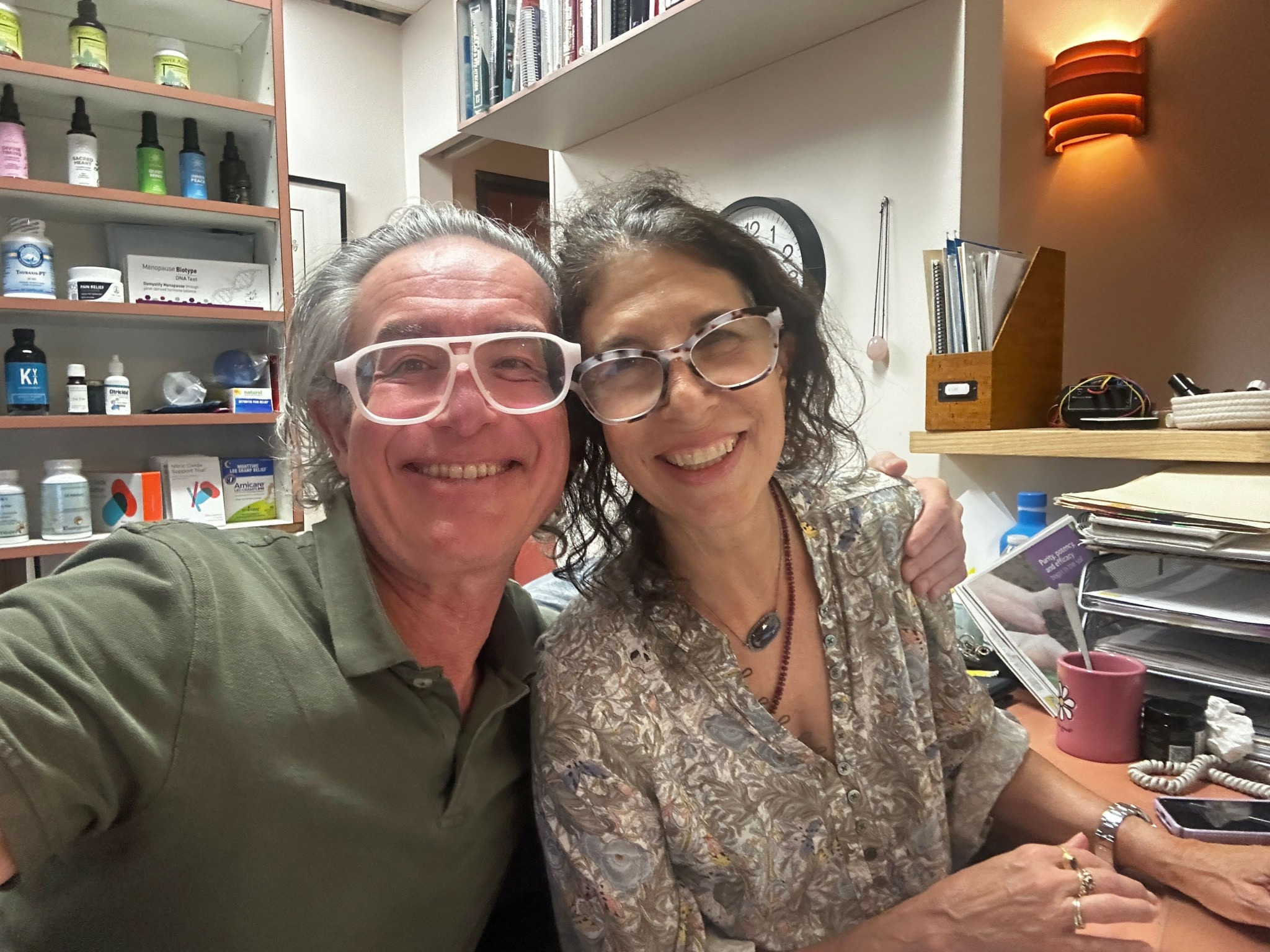 Two people smiling, one wearing glasses and a patterned top, the other in a green shirt, in an indoor setting.