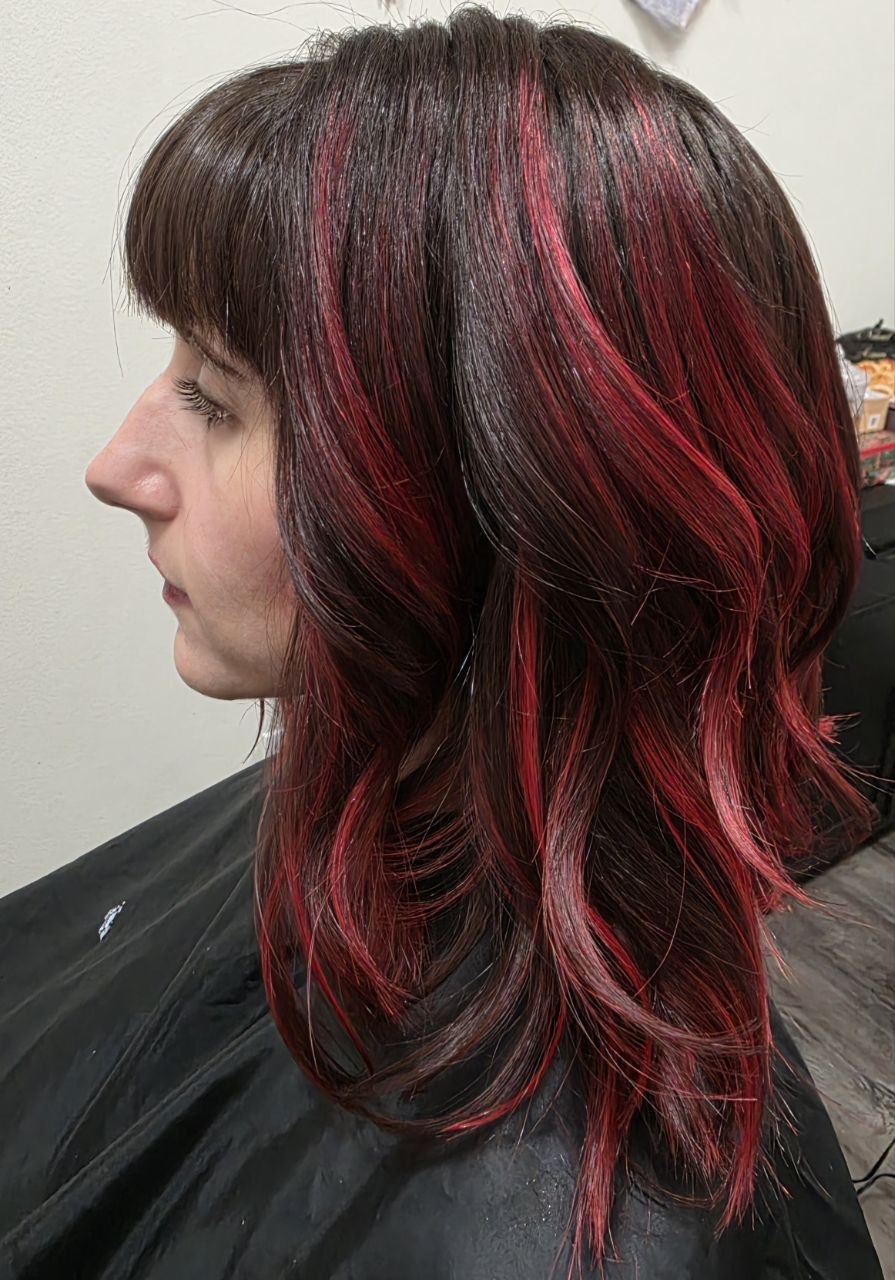 Side view of a woman with shoulder-length dark hair and red highlights, styled in loose waves.