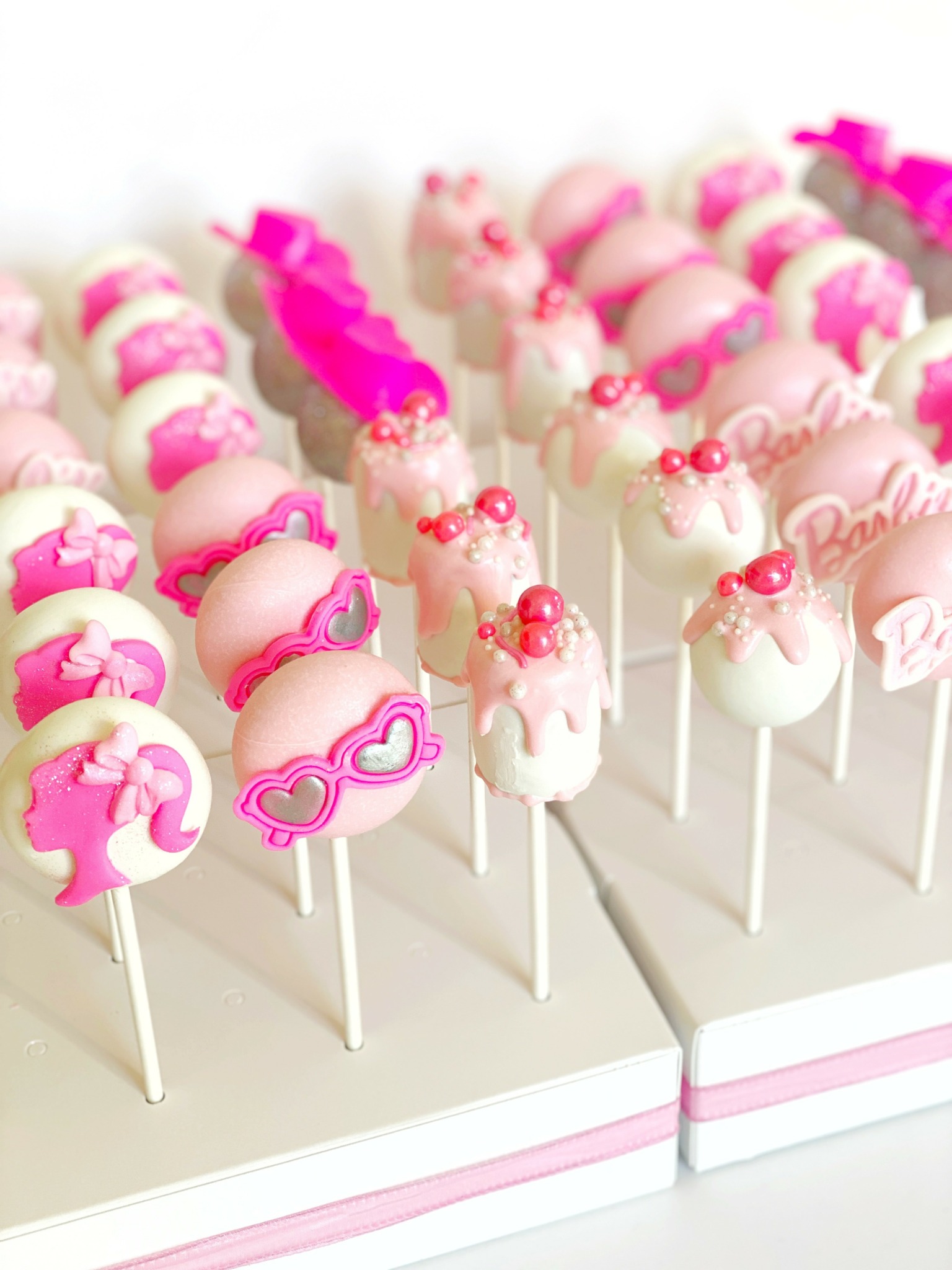 Pink and white cake pops decorated with bows, glasses, and pearls on sticks, arranged on a white tray.
