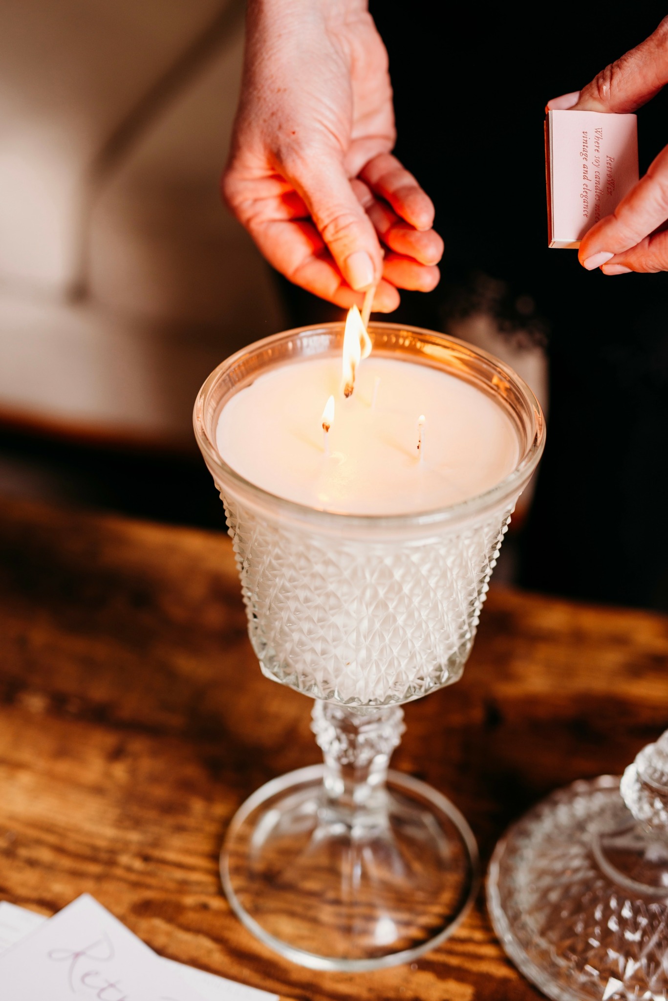Person lighting a candle in a decorative glass holder on a wooden table.