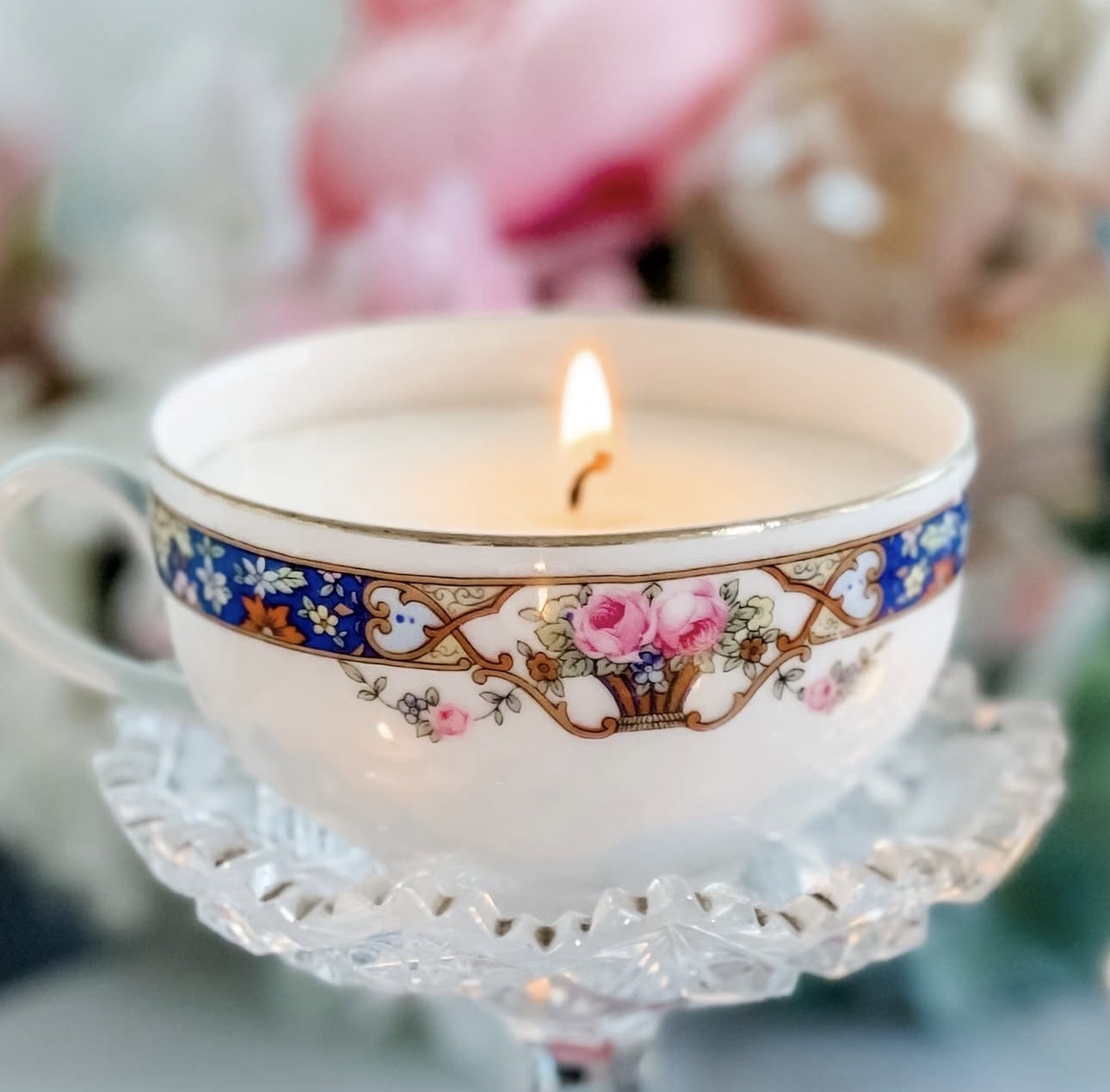 Decorative teacup with a lit candle inside, placed on a glass stand with blurred floral background.