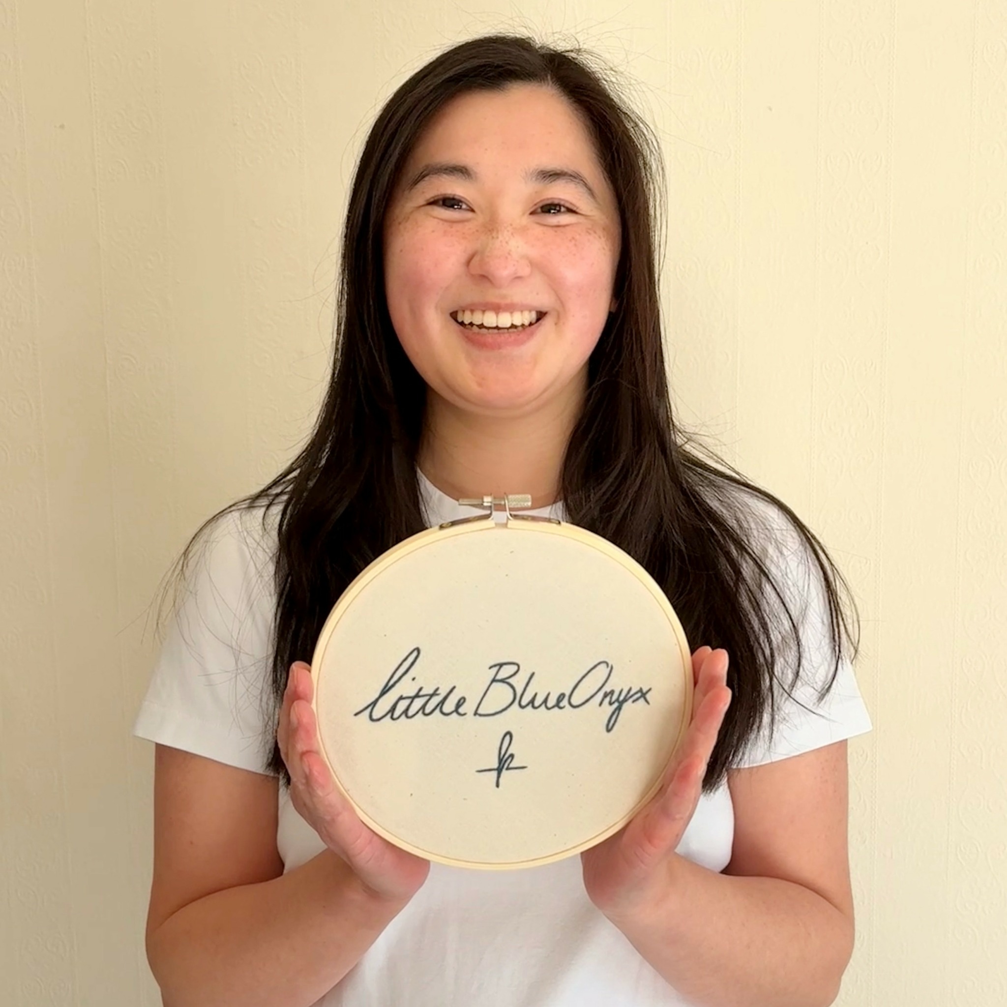 Young woman with long dark hair smiling, holding a round embroidery hoop with handwritten text, standing against a plain wall.