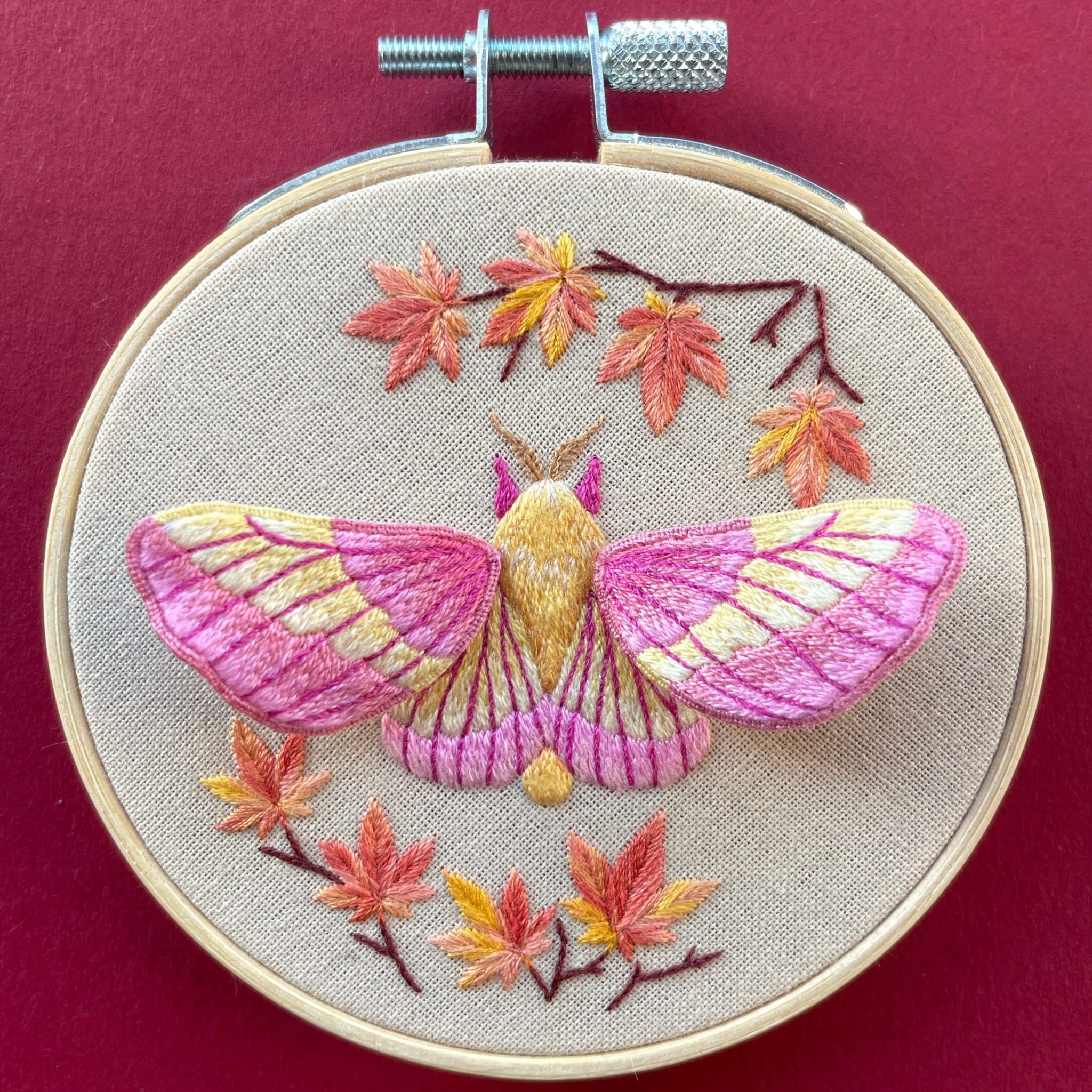 Embroidery of a pink and yellow butterfly with autumn leaves on beige fabric, in a wooden hoop.