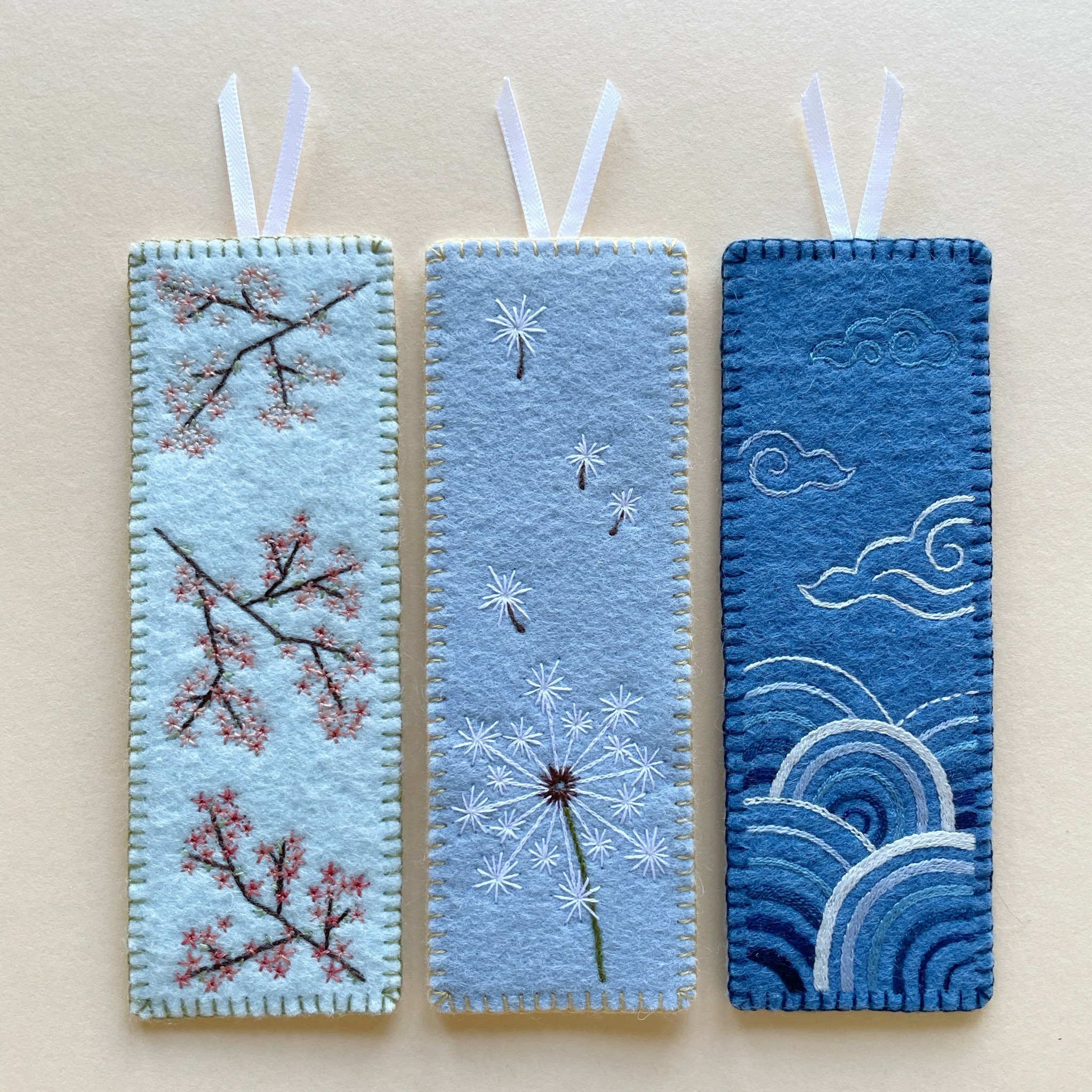 Three embroidered fabric bookmarks with sky and cloud designs, hanging on white ribbons against a light background.
