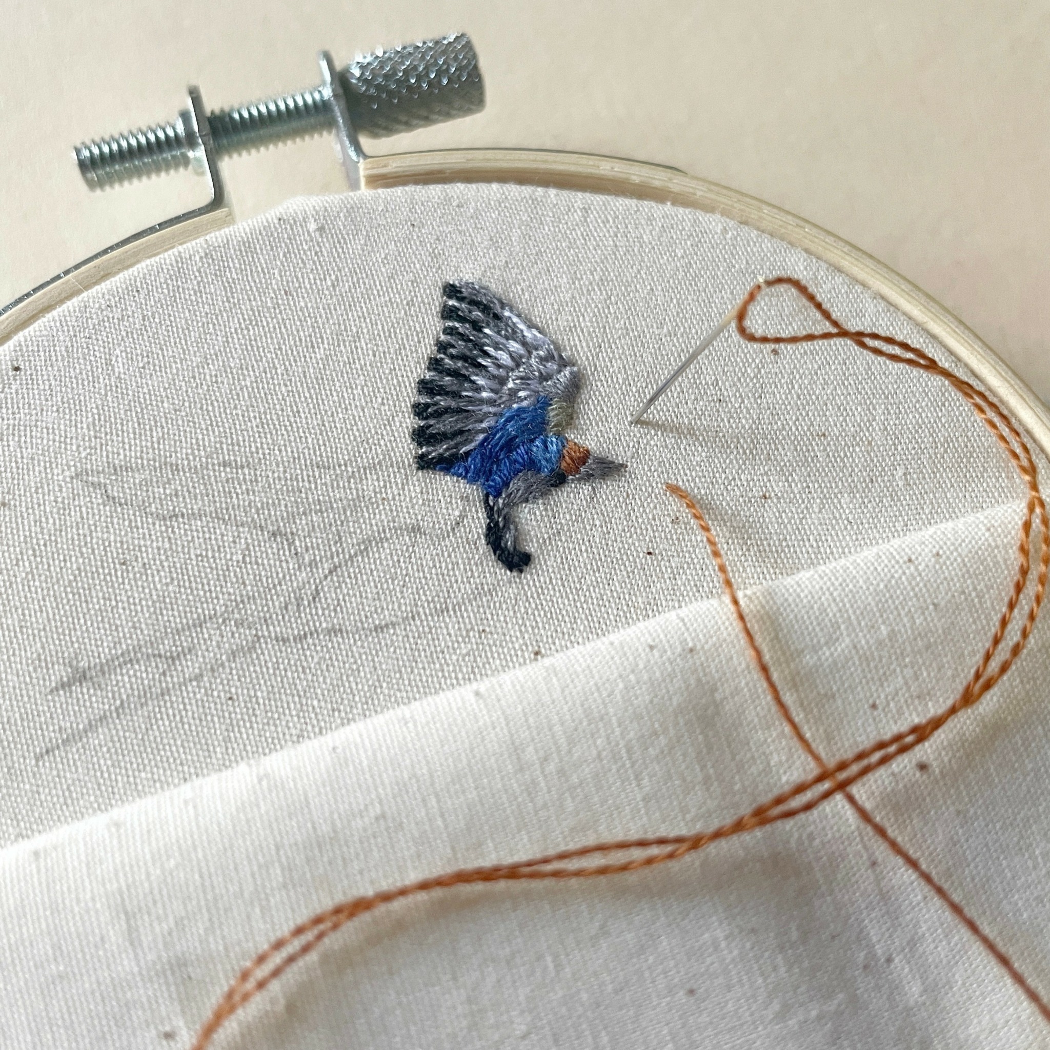 Embroidery hoop with a partially stitched bird and thread on fabric, with a needle and thread nearby.