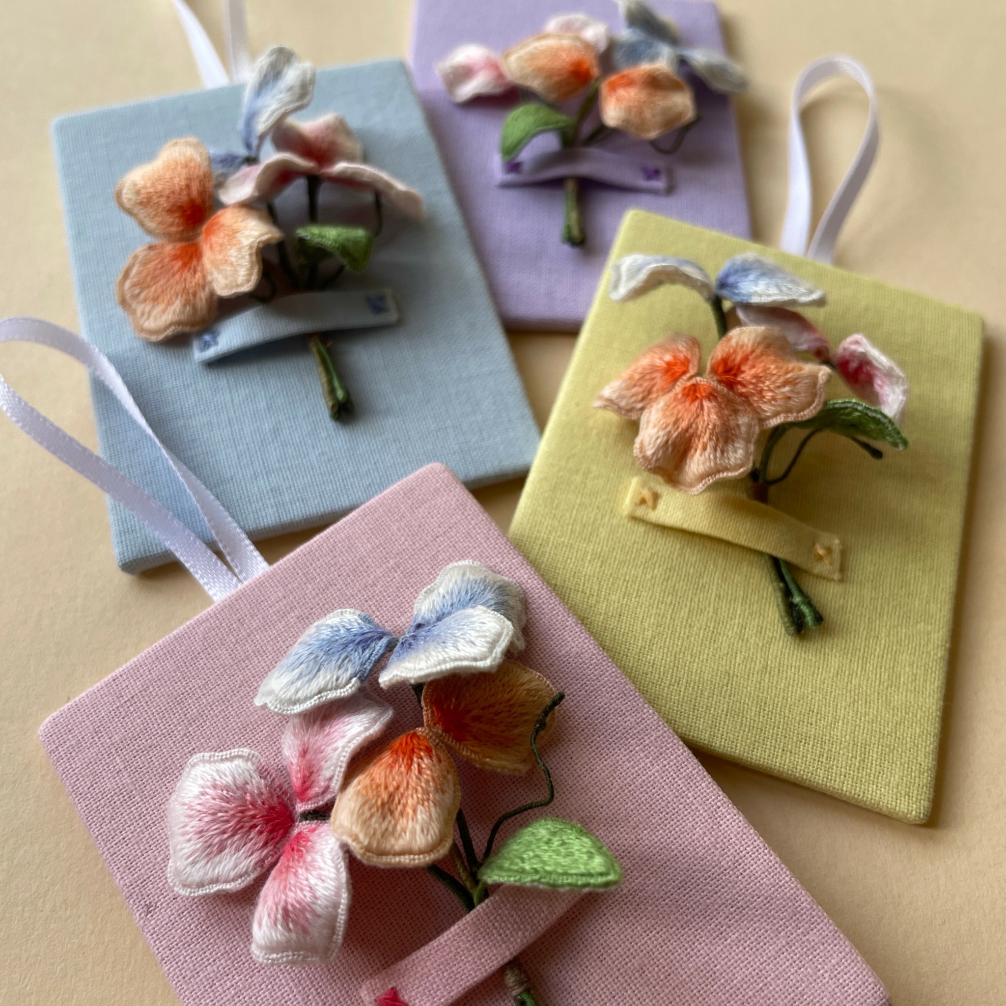 Four fabric-covered notebooks with embroidered flower embellishments in pastel colors, arranged on a flat surface.