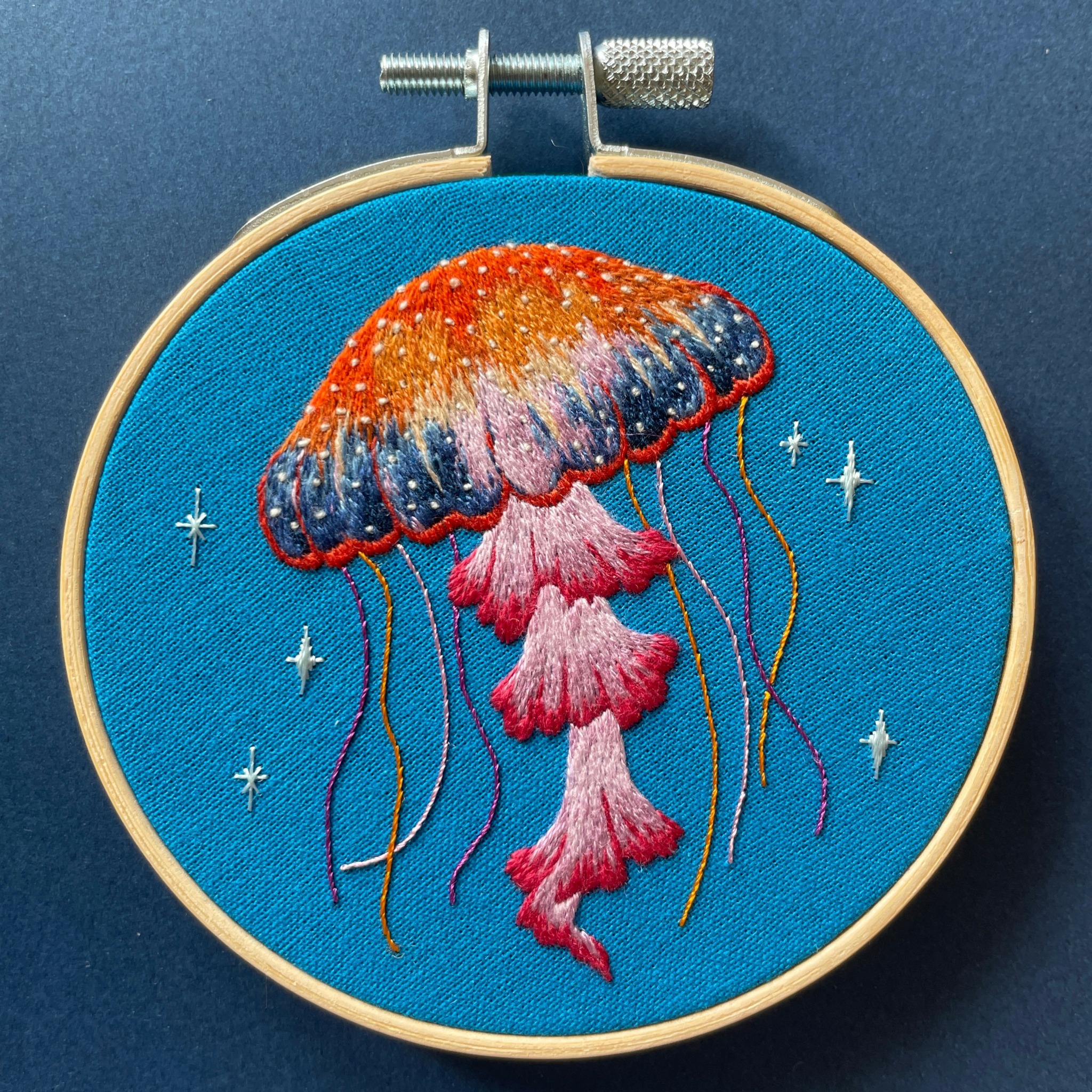 Embroidery of a jellyfish with orange, pink, and purple colors on blue fabric background.