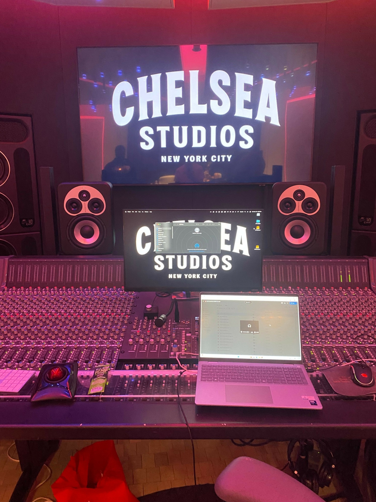 Music studio setup with mixing console, speakers, and a laptop on a desk, with a large screen displaying 'Chelsea Studios'.