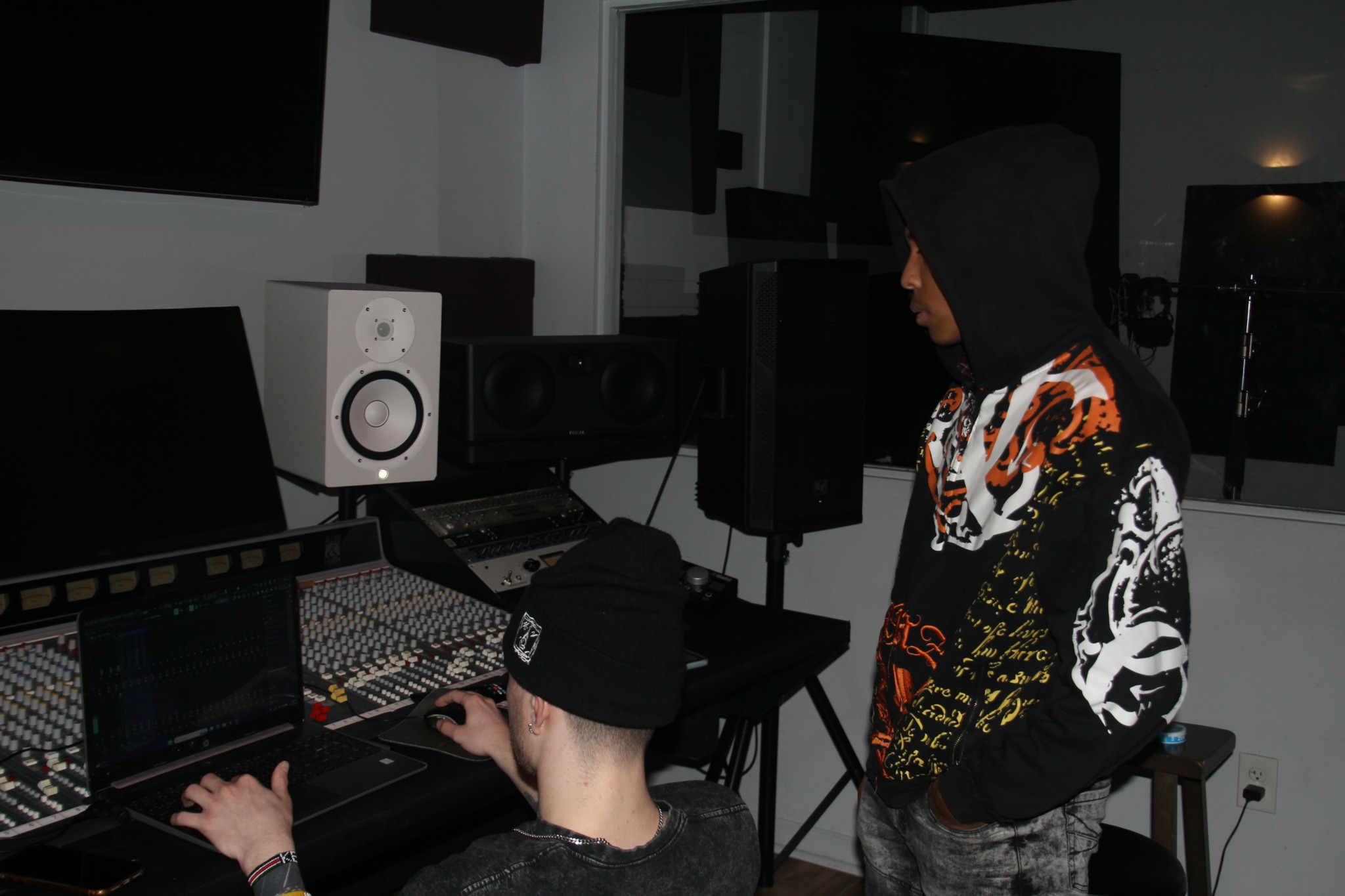 Two people in a music studio, one working on a computer and the other standing nearby, wearing a hoodie and patterned jacket.
