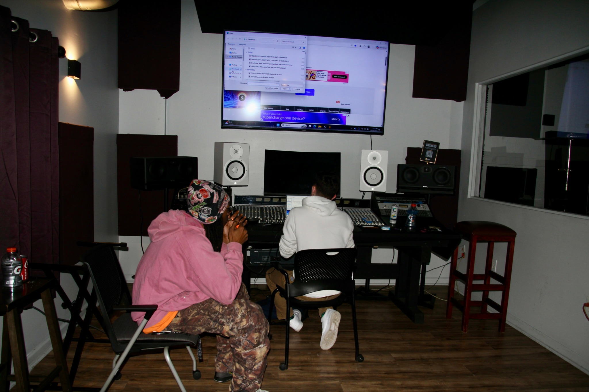Two people sit at a mixing console in a recording studio, facing a large screen displaying a digital audio workstation.