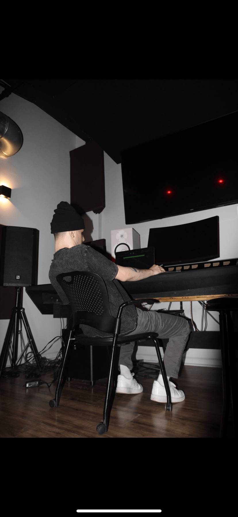Person sitting at a music studio desk with computer monitors and audio equipment, wearing a beanie and dark clothing.