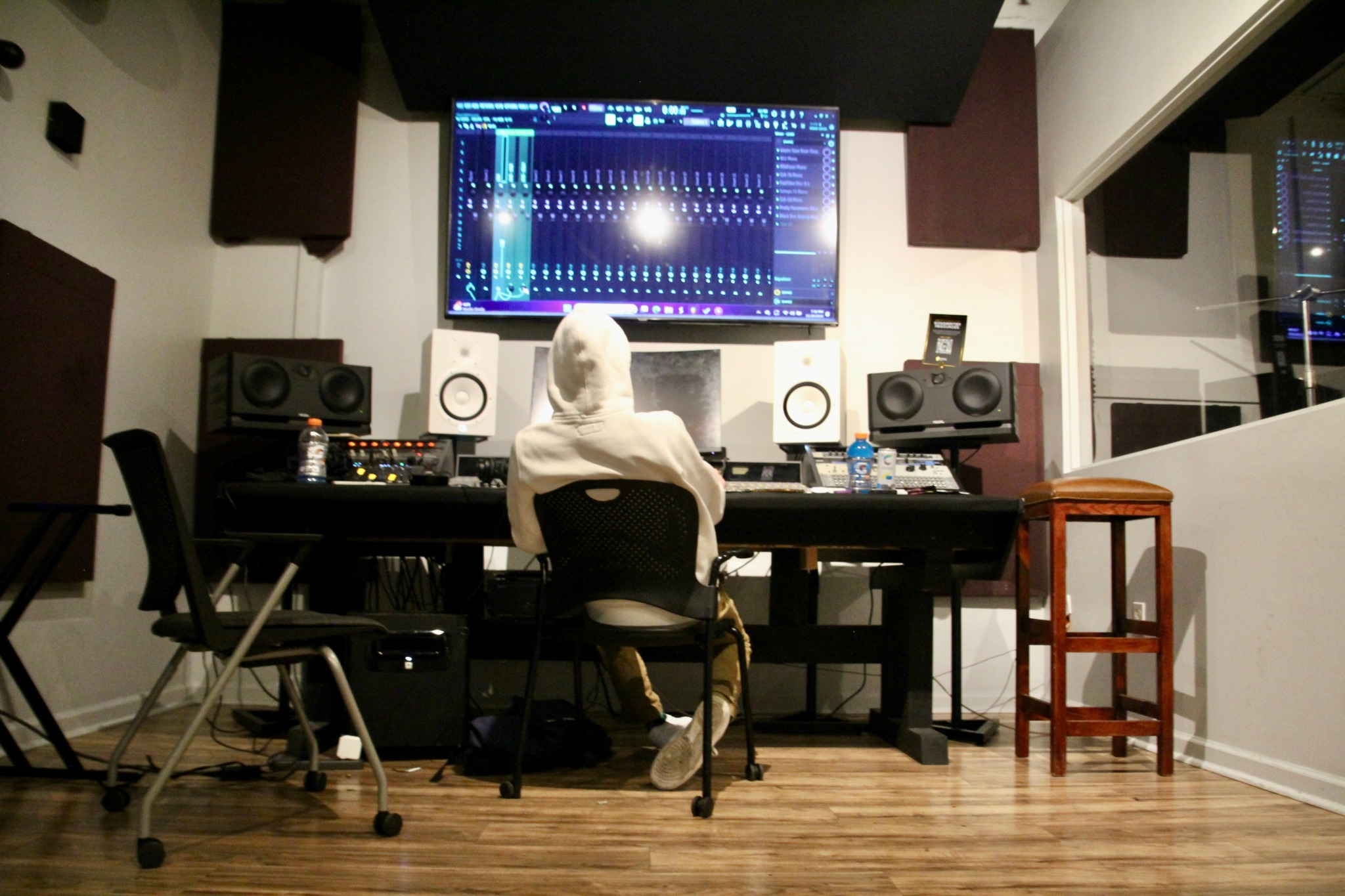 Person wearing a hoodie working at a music production studio with speakers and a large screen displaying audio software.