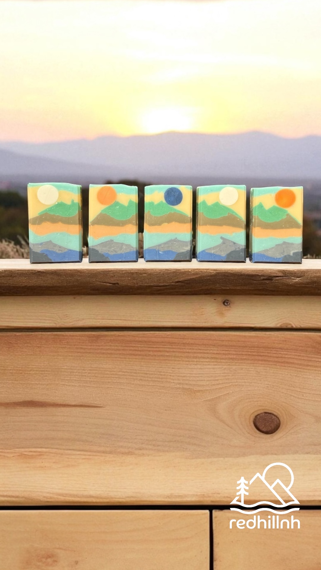 Five small paintings of landscapes with mountains, sky, and water, placed on a wooden surface with a sunset background.
