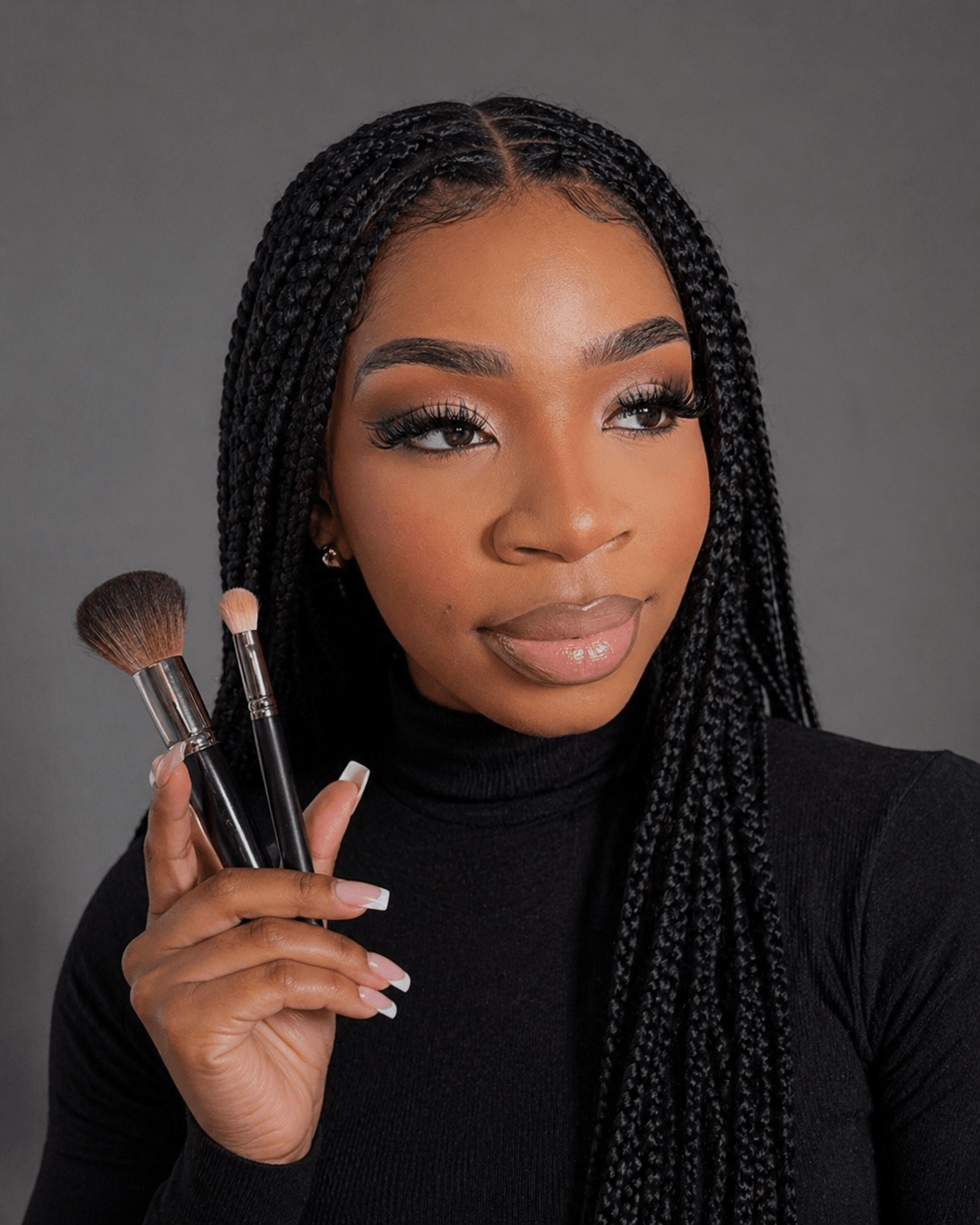 Woman with braided hair holding makeup brushes, wearing a black top, looking to the side.