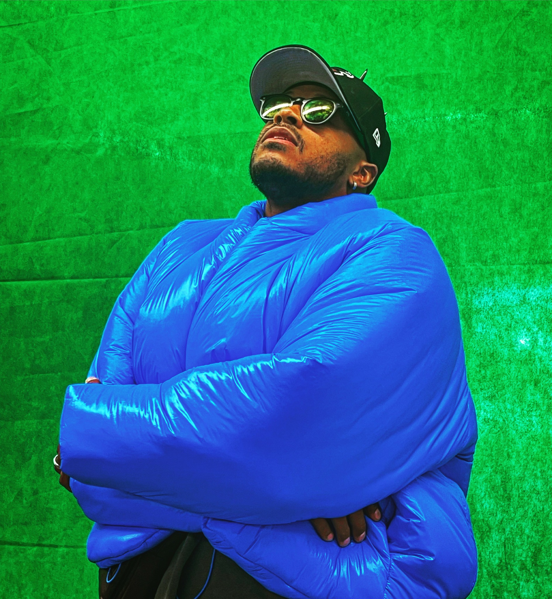 Person wearing sunglasses, cap, and a bright blue puffer jacket, standing against a green background.