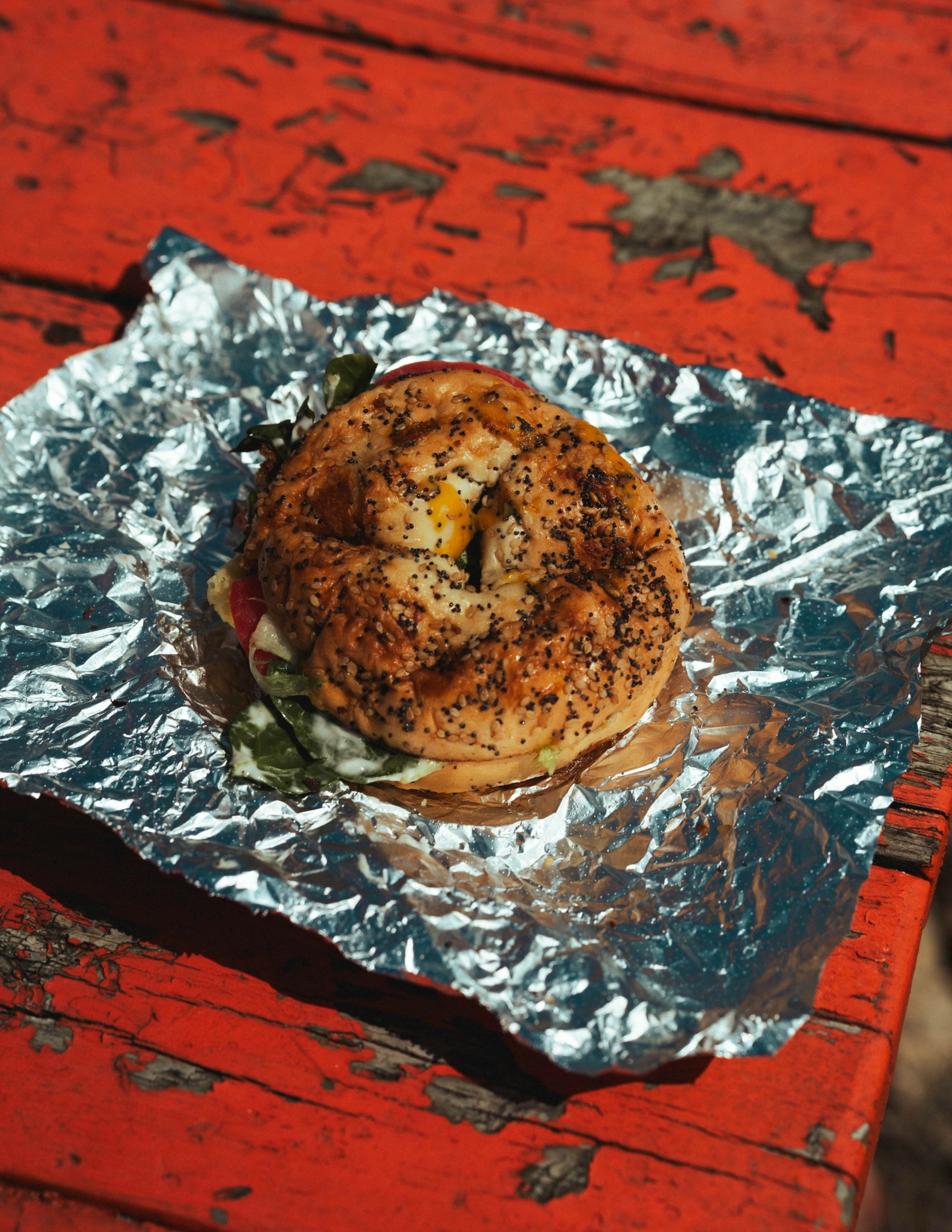 Bagel with toppings on crumpled aluminum foil on red wooden surface.