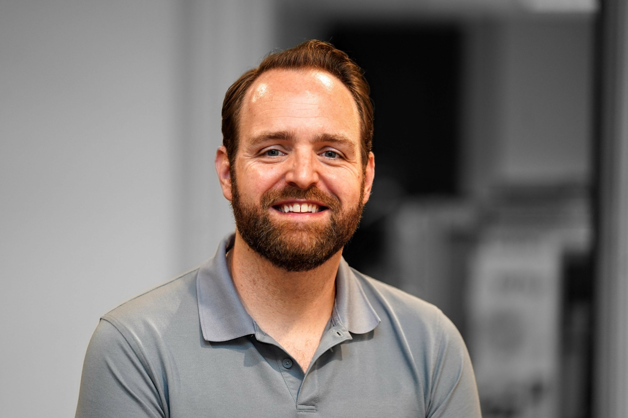 Man with beard smiling, wearing a gray polo shirt, in an indoor setting.