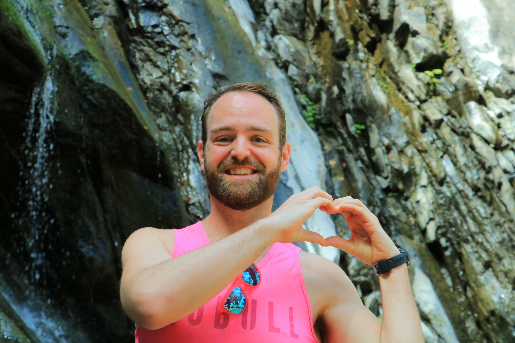 Man with a beard wearing a pink tank top making a heart shape with his hands outdoors.