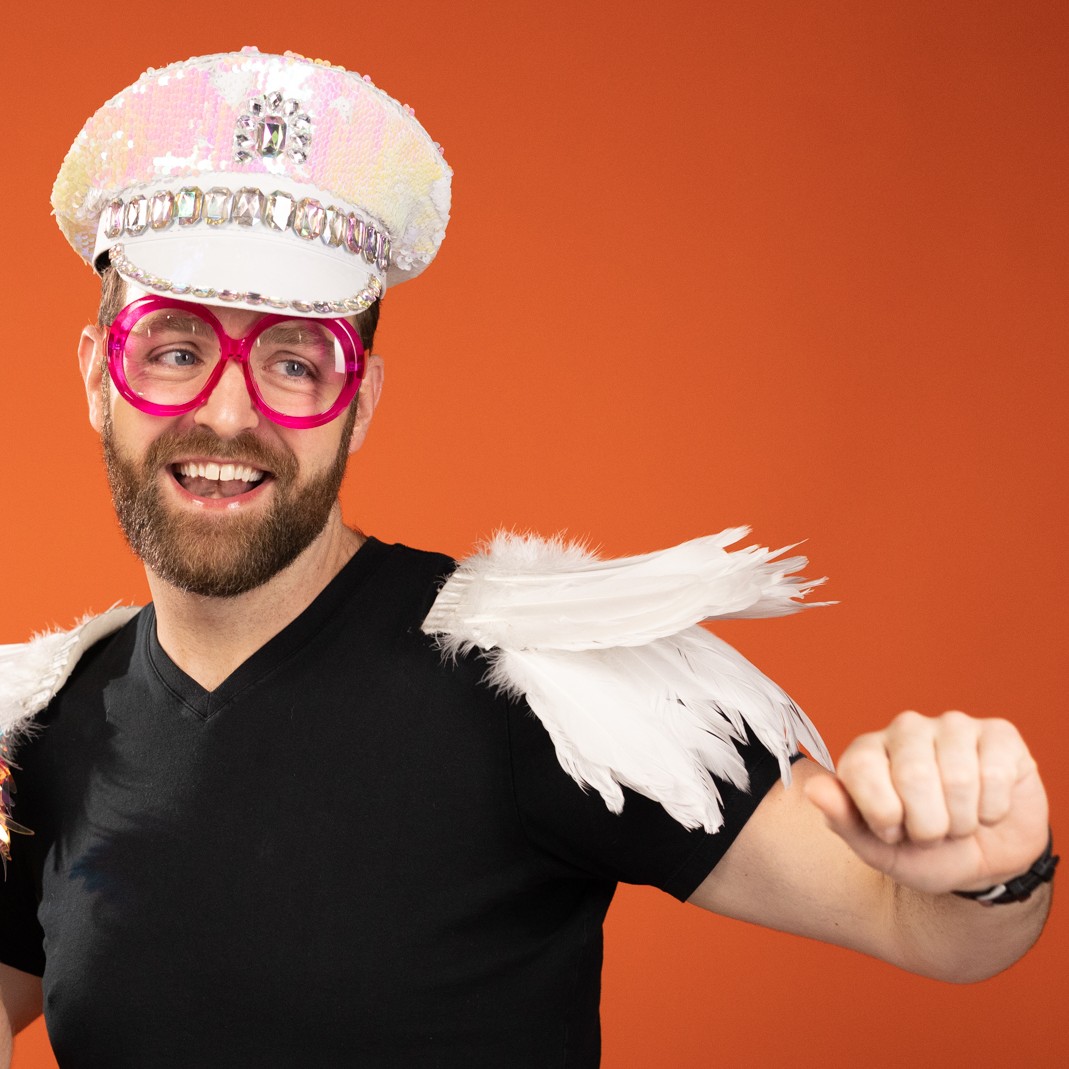 Person wearing a sequined hat, pink glasses, and a costume with white feathers on shoulders, smiling and flexing arm.
