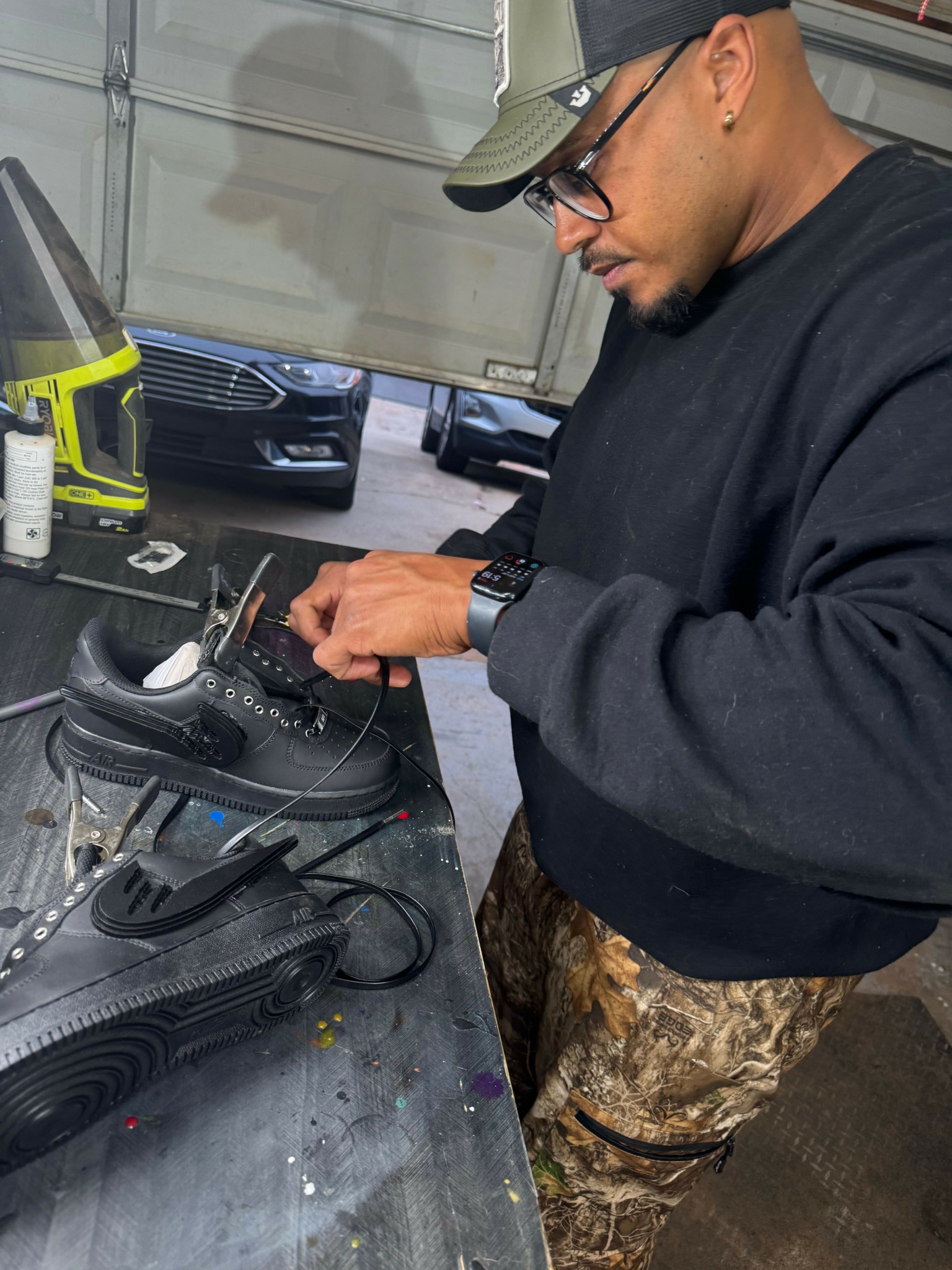 Person wearing glasses, black hoodie, and camouflage pants working with electronic equipment on a table.