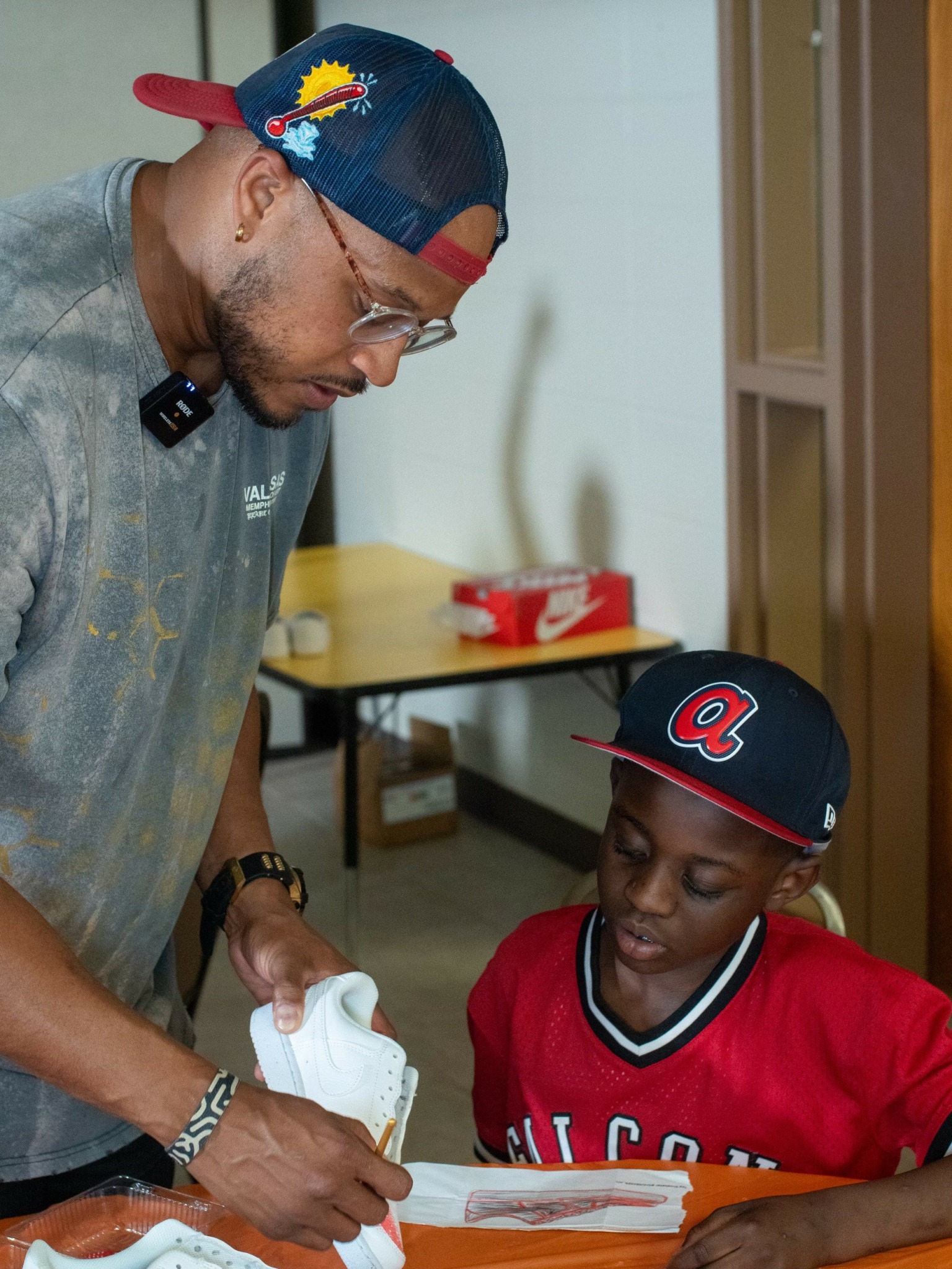 Man and boy looking at a paper on a table indoors, man holding a pen, boy wearing a cap and red shirt.