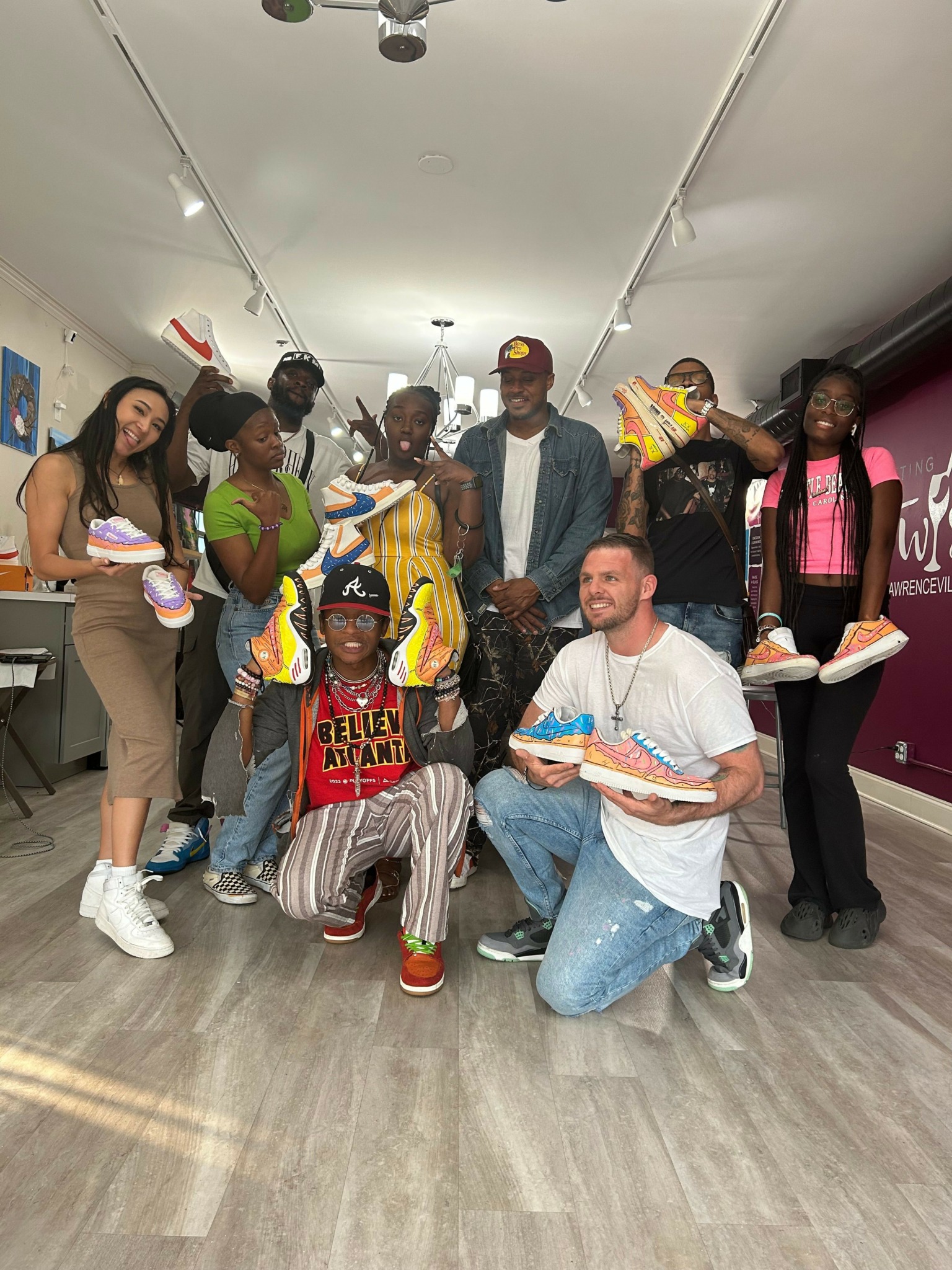 Group of people standing and sitting in a hallway, some holding food and drinks, with a luggage cart in the background.