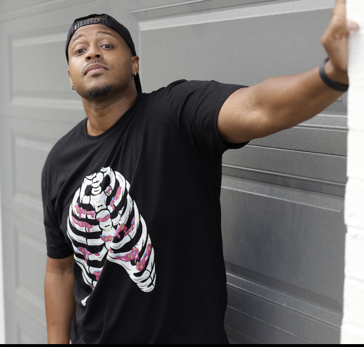 Young man wearing a black T-shirt with a colorful zebra print, standing against a gray wall, looking at the camera.