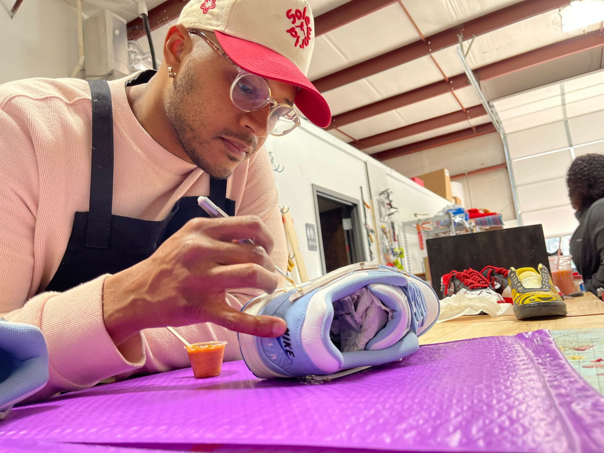 Person wearing glasses, pink cap, and apron, painting a marble-like object on a purple mat in a workshop.