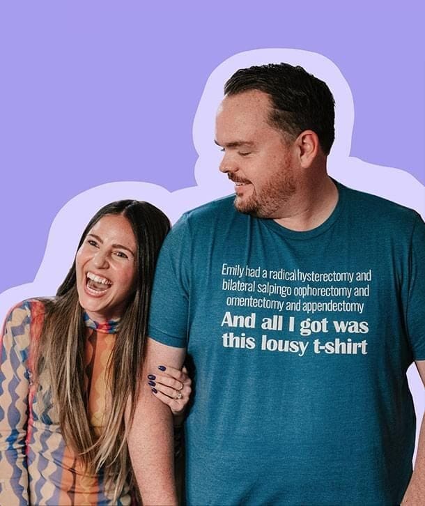 Two people smiling and laughing, one wearing a blue t-shirt with white text, the other with long hair and a patterned top.