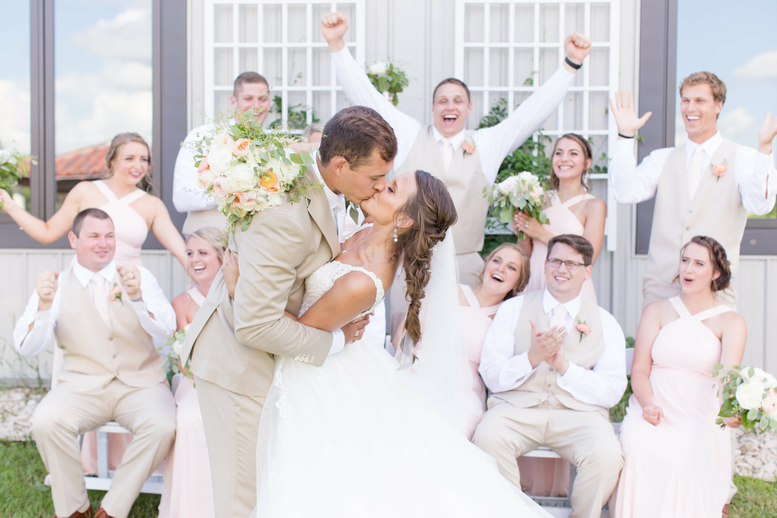 Bride and groom kissing outdoors with wedding party celebrating behind them, holding bouquets and smiling.