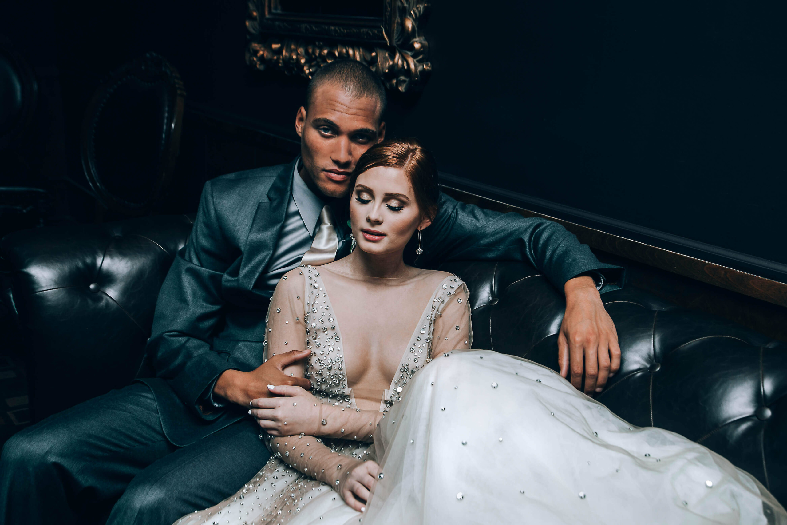 Man and woman sitting on a black leather couch in a dark room, woman in a white dress, man in a suit.