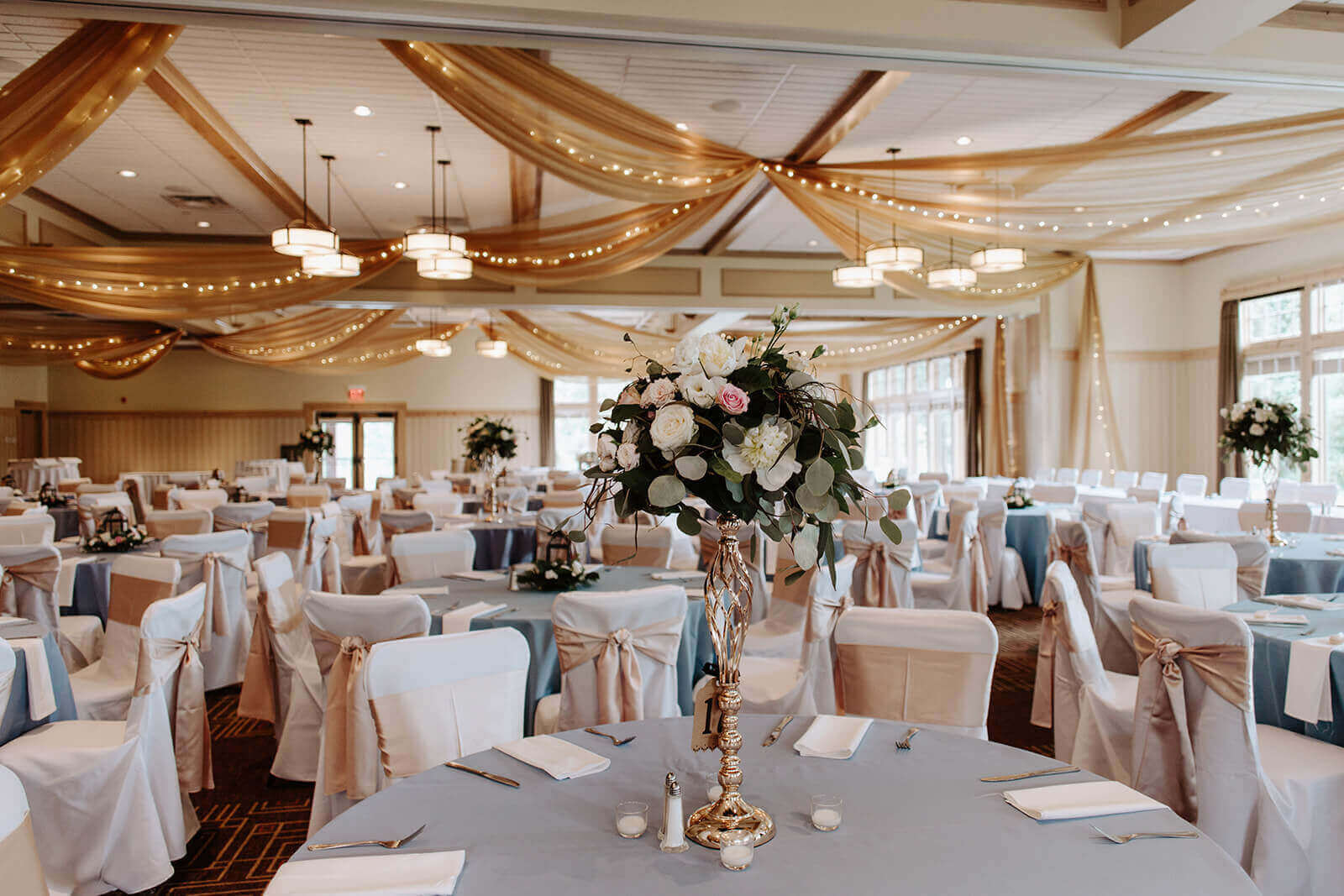 Elegant banquet hall with round tables, white chairs, floral centerpieces, and draped ceiling decorations.